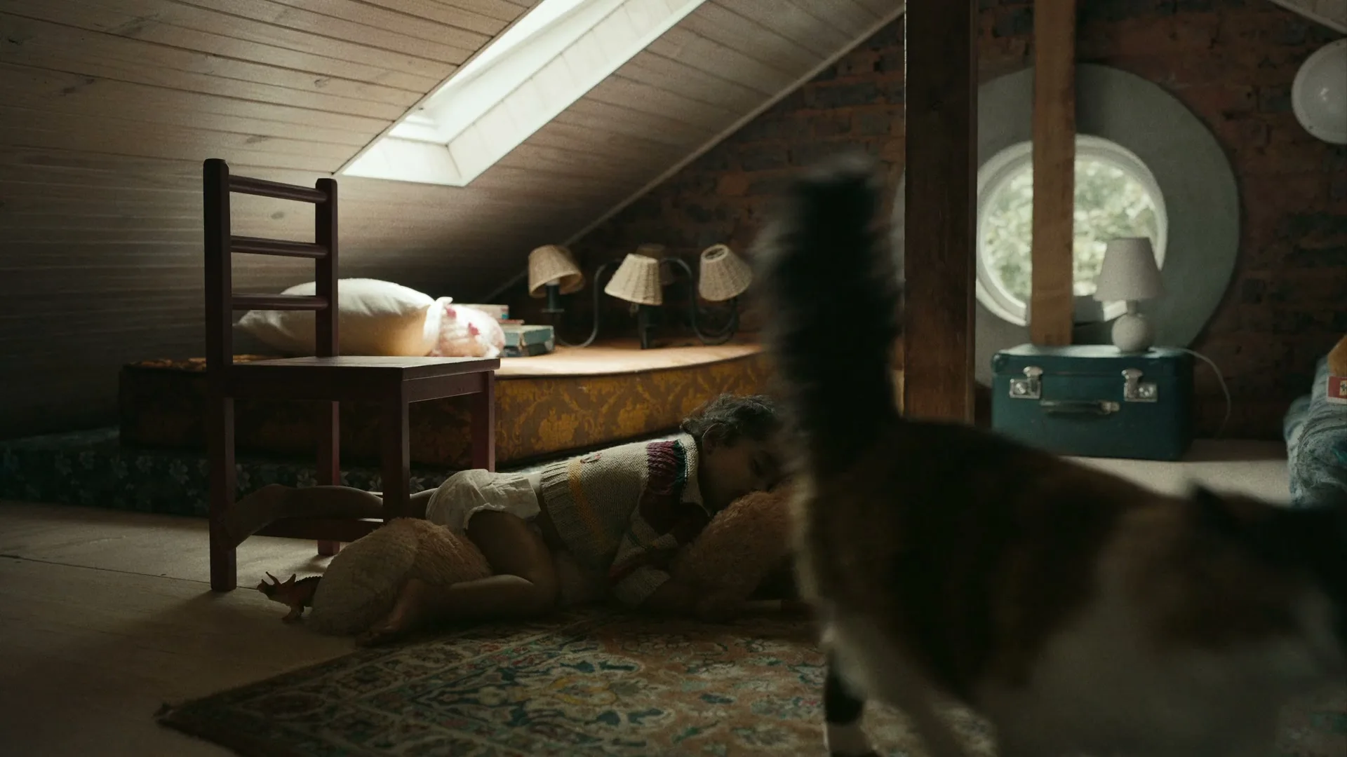 A young child sleeps on the floor, hugging a teddy bear, as a cat walks past in a cozy attic room.