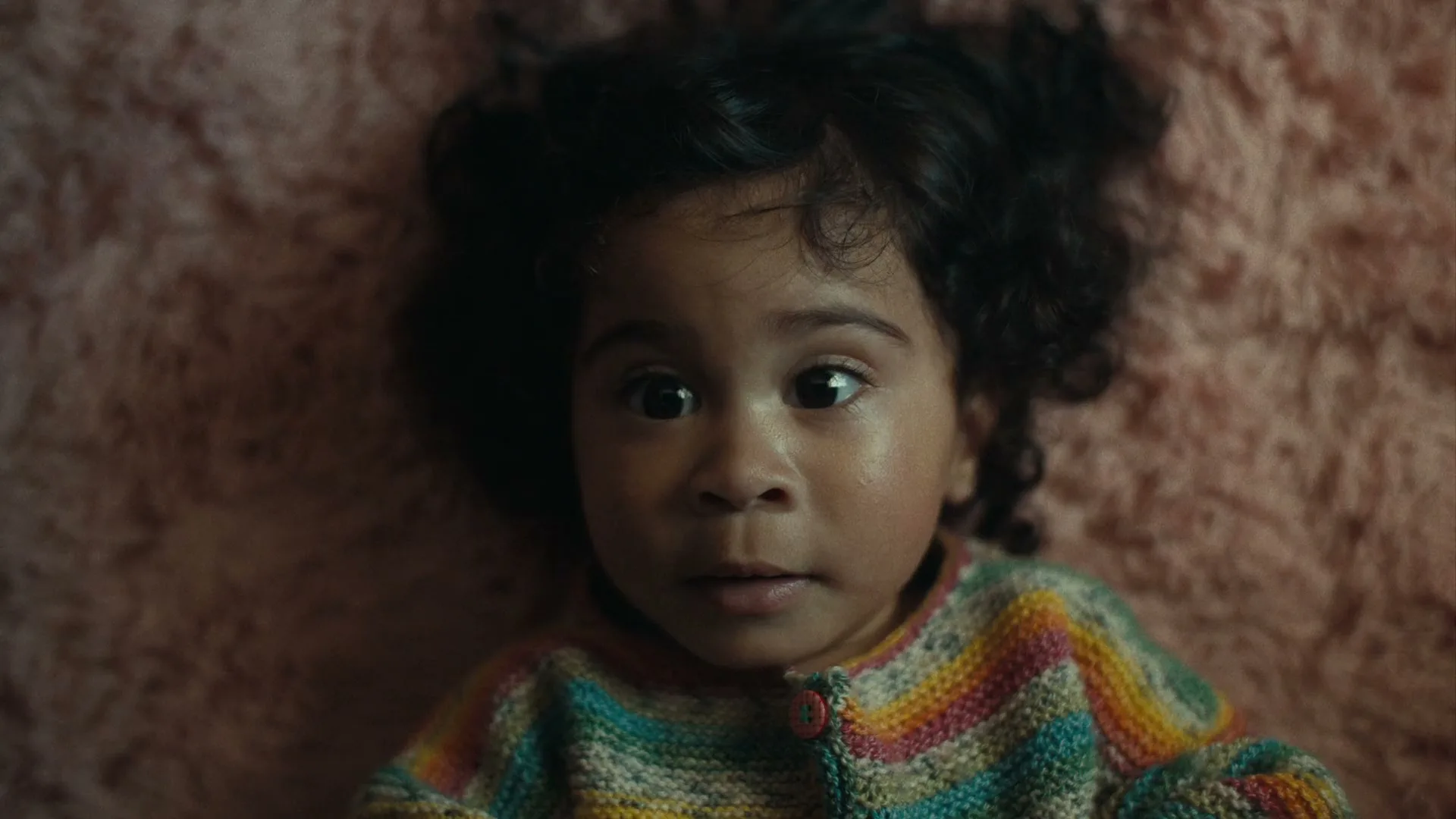 A baby with dark curly hair and a colorful sweater lies on a pink fuzzy blanket, looking up with wide eyes.