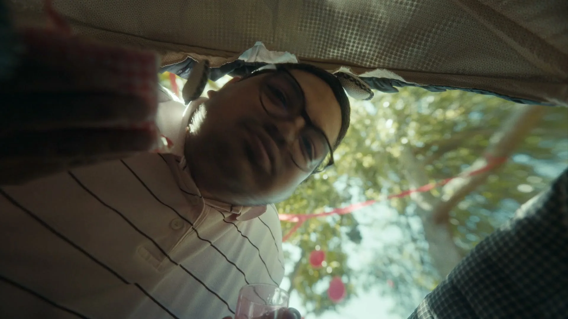 A man with glasses and a striped shirt looks down into a baby's crib, holding a drink, at an outdoor party.