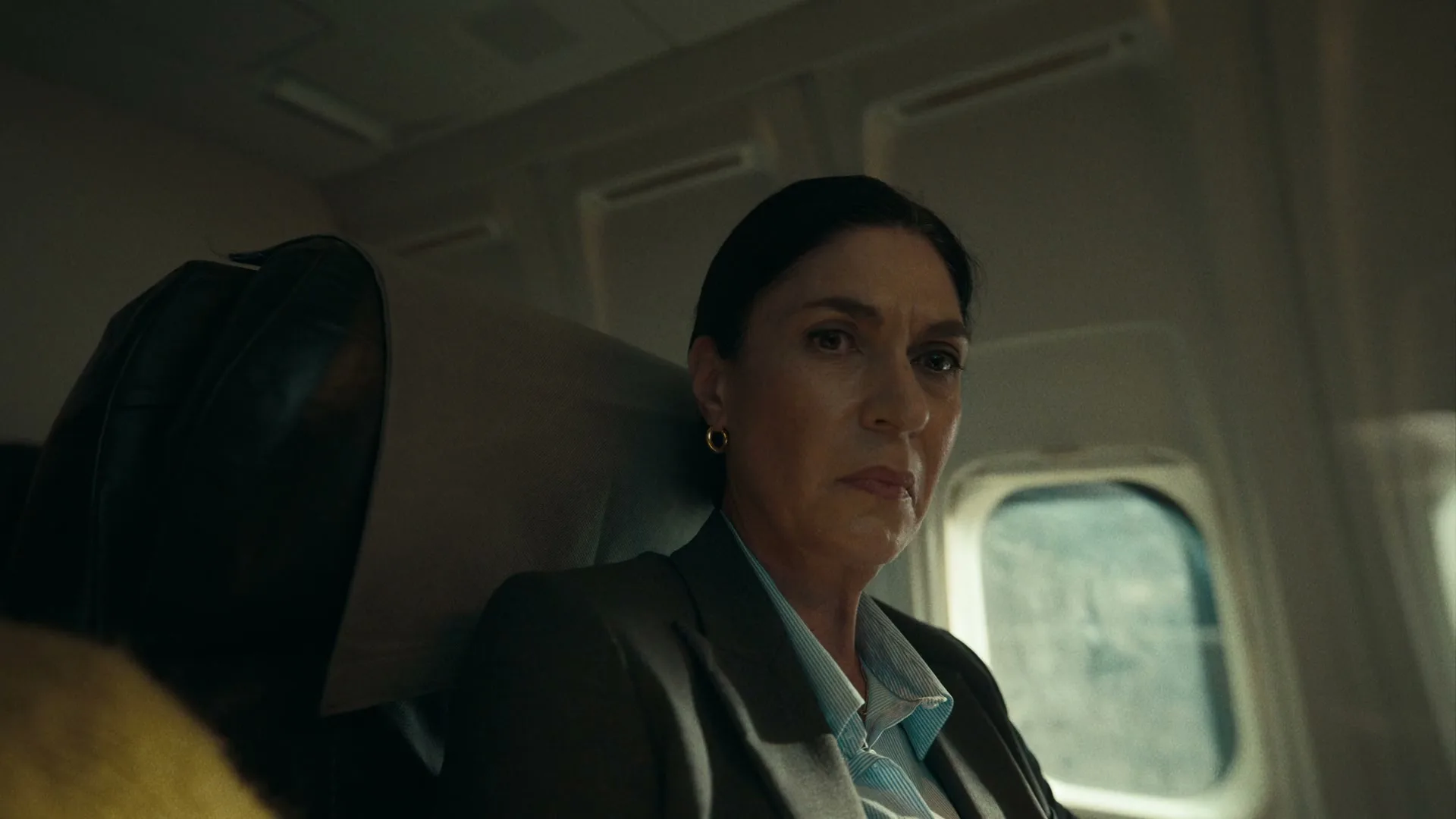 A woman with dark hair in a business suit sits in an airplane seat, looking forward with a concerned expression, then turns her head to the right.