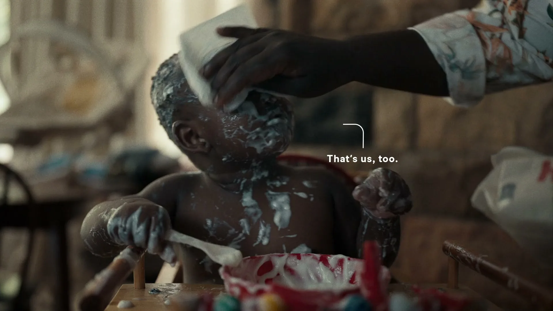 A baby covered in food sits in a high chair as an adult hand wipes their face with a tissue in a home.