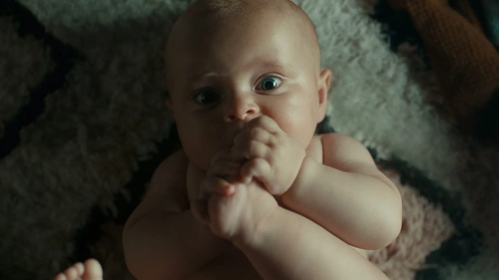 A baby lies on a patterned rug, looking up at the camera with wide eyes, bringing its foot to its mouth.