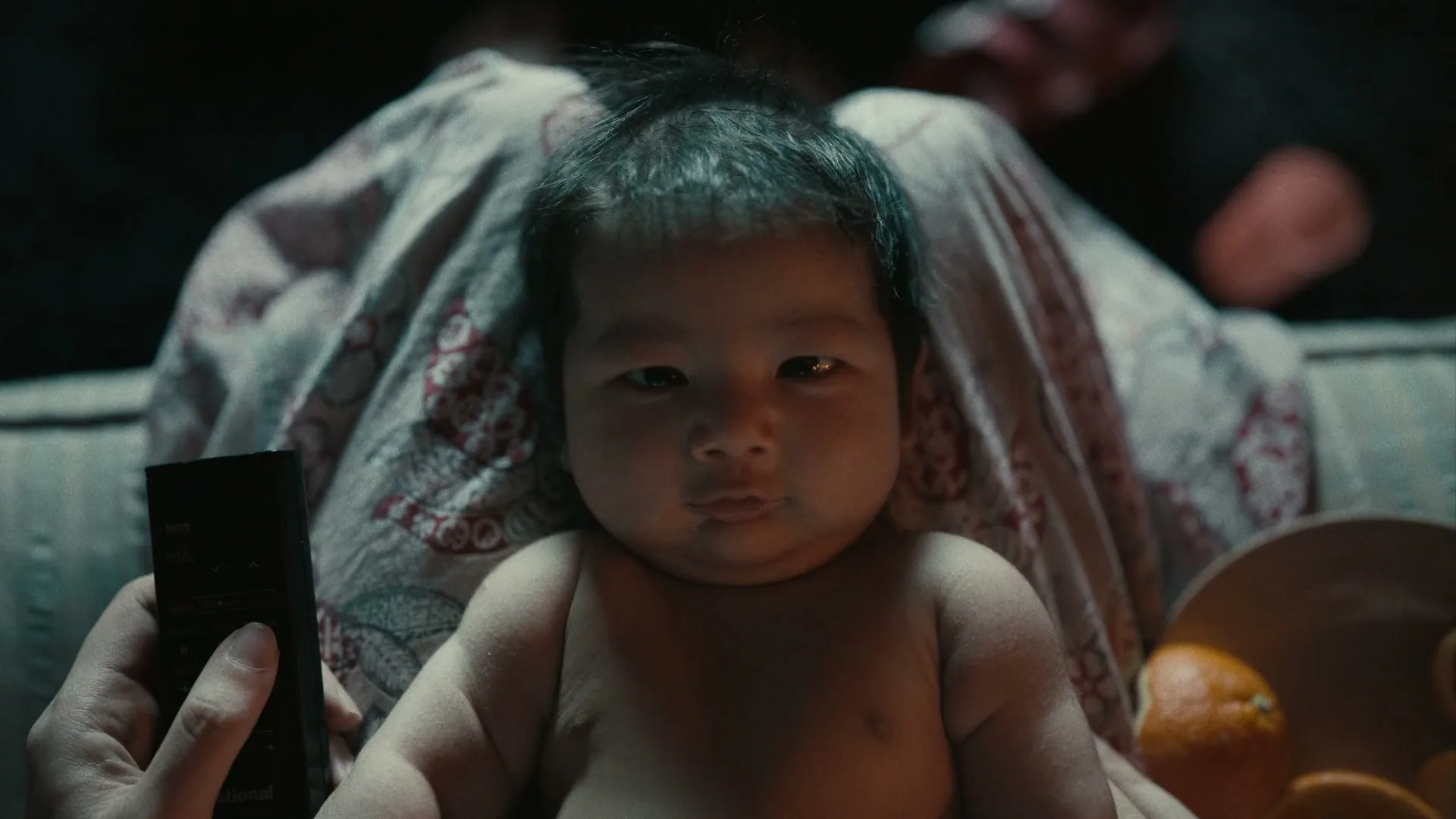 A baby with dark hair sits on an adult's lap, looking directly at the camera, with a hand holding a remote control nearby.