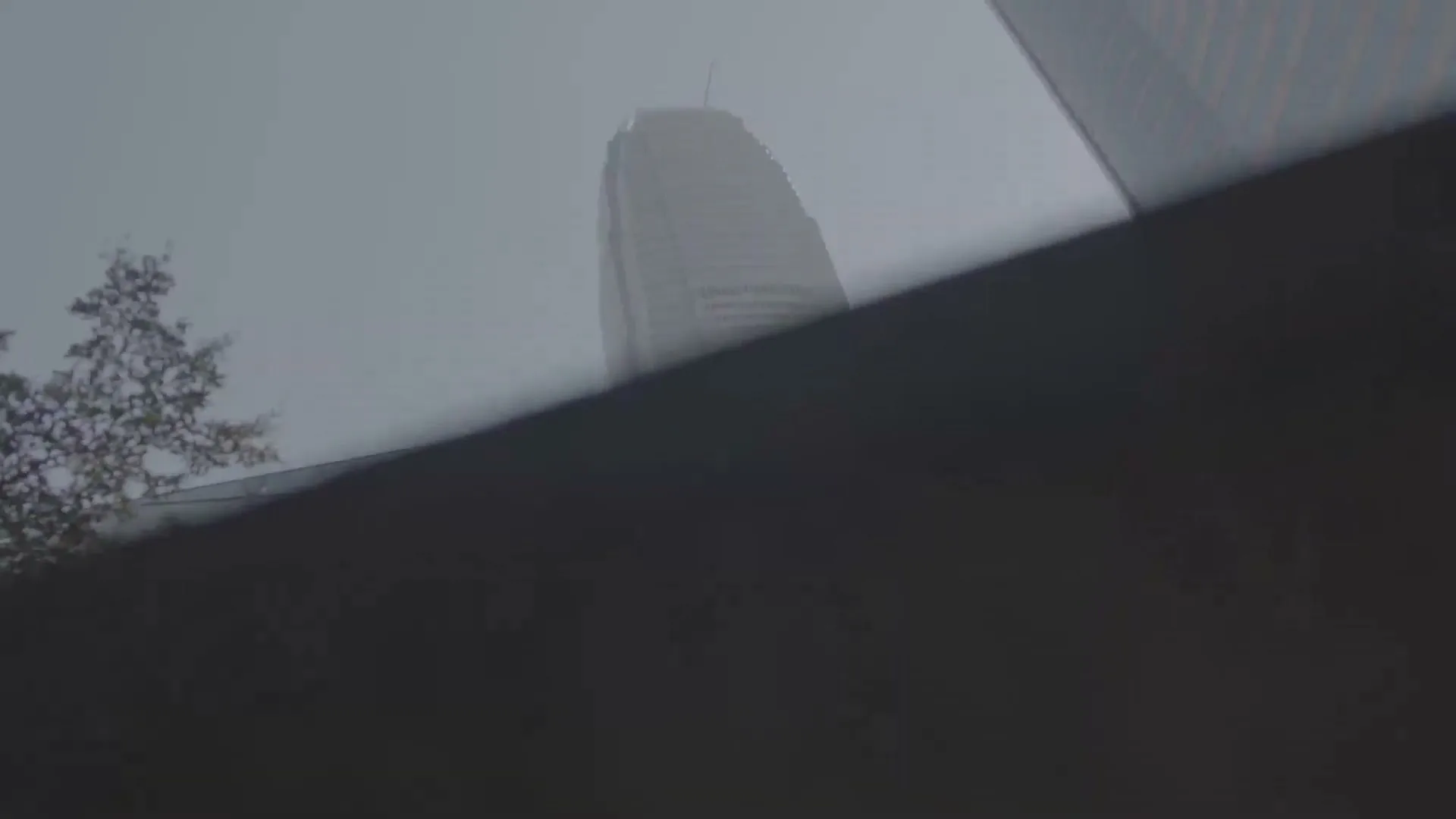A low-angle shot reveals a modern skyscraper shrouded in mist, with a pedestrian bridge and trees in the foreground, creating a sense of urban mystery.