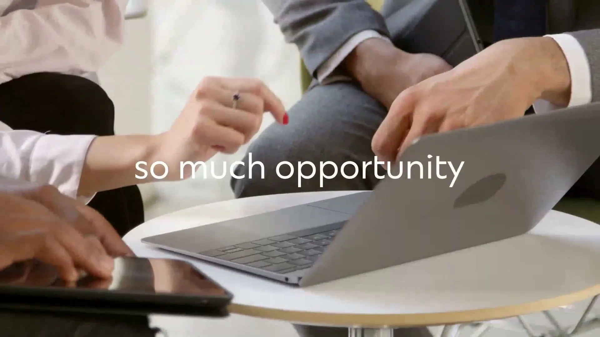 Three business people, two men and one woman, are gathered around a small table, looking at a laptop and a tablet in an office.