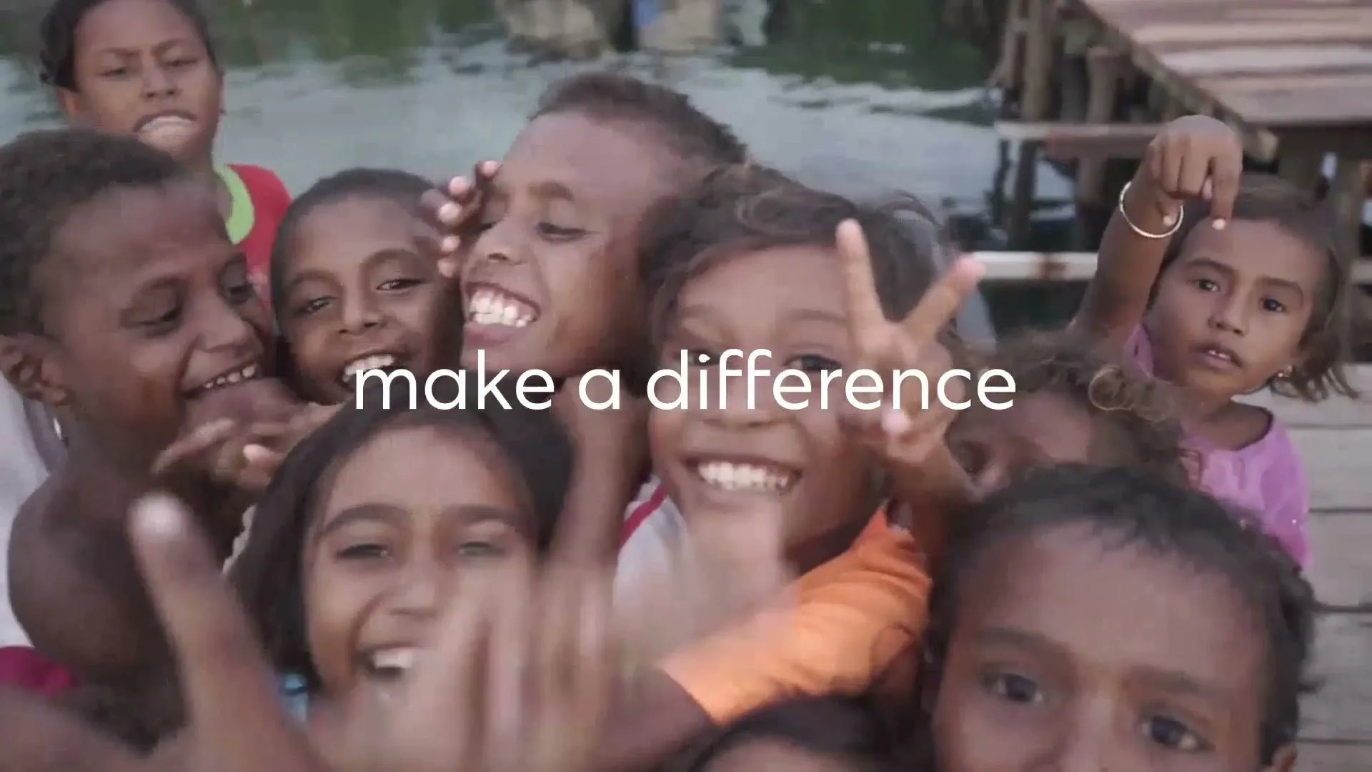 A group of diverse children, some smiling and making peace signs, are gathered closely together on a wooden dock over water.