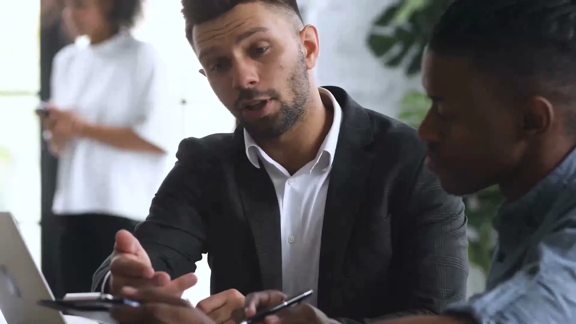 Two male colleagues, one white and one Black, discuss work at a laptop in a modern office, with a female colleague blurred in the background.
