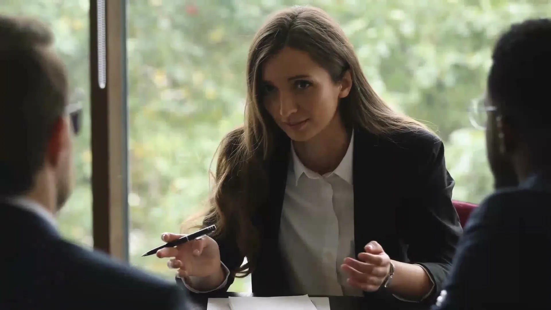 A young businesswoman with long brown hair, wearing a black suit, gestures with her hands while speaking to two men in a modern office with a large window.