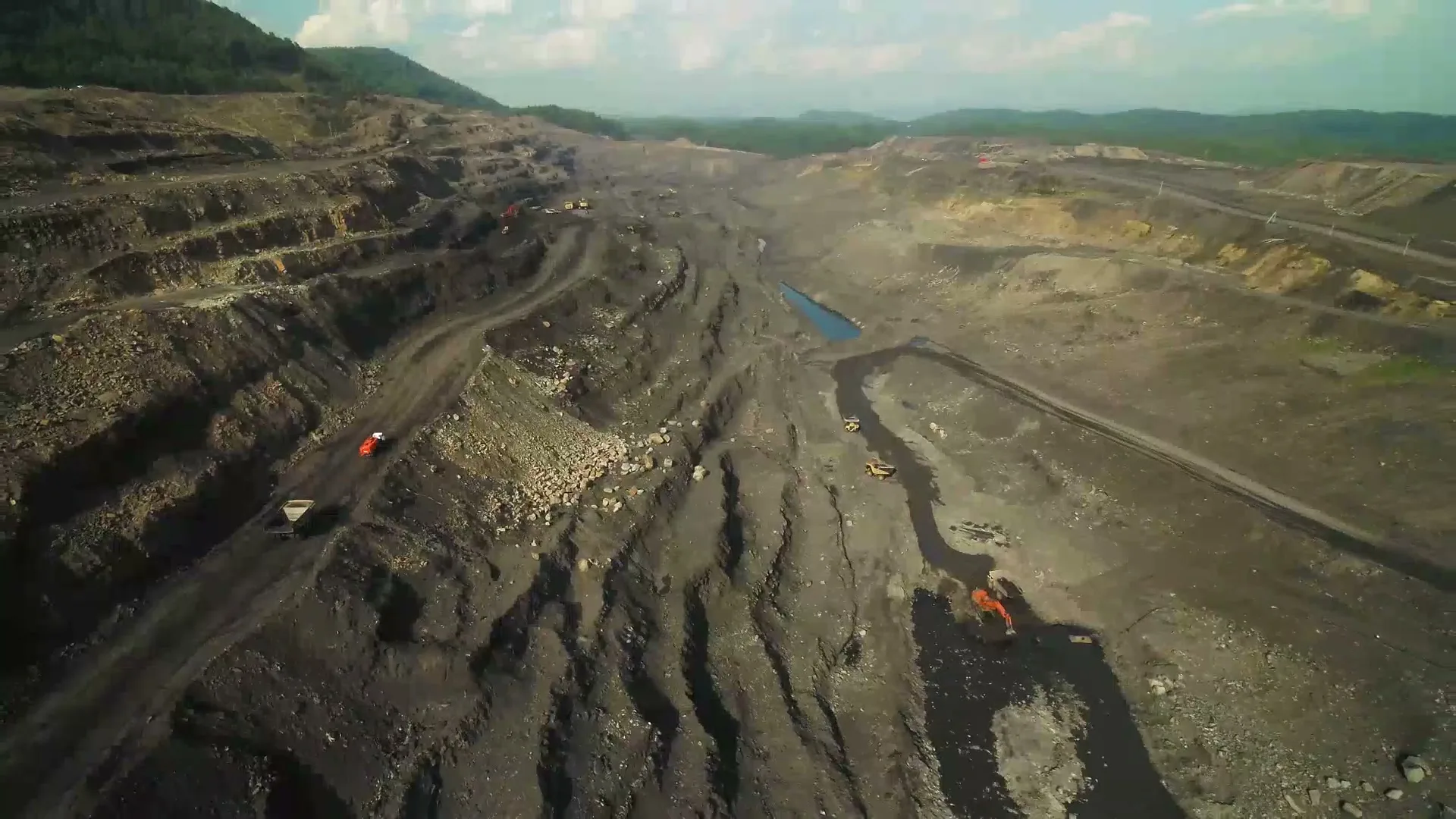 An aerial view shows a vast open-pit coal mine with heavy machinery operating on the dark, excavated terrain under a cloudy sky.