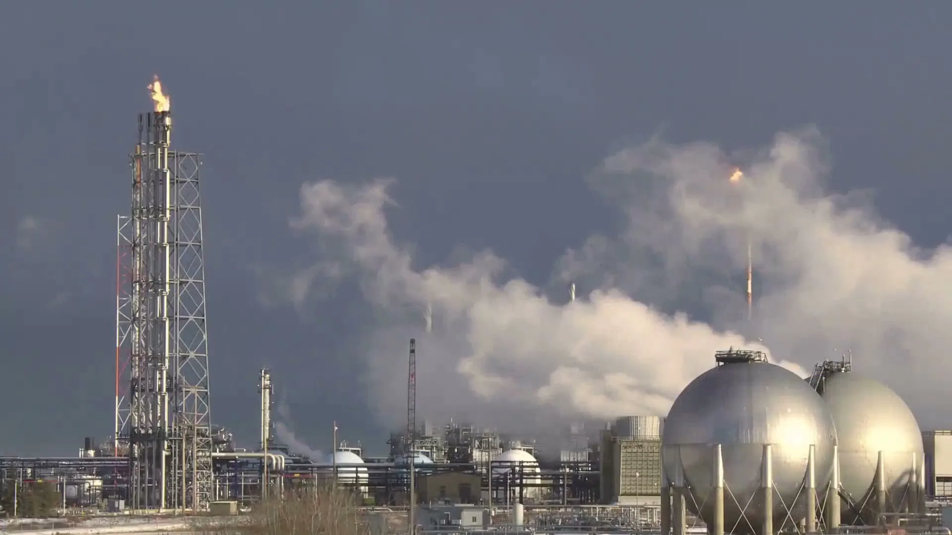 A wide shot of an industrial oil refinery with tall towers, spherical tanks, and plumes of smoke and fire against a dark sky.