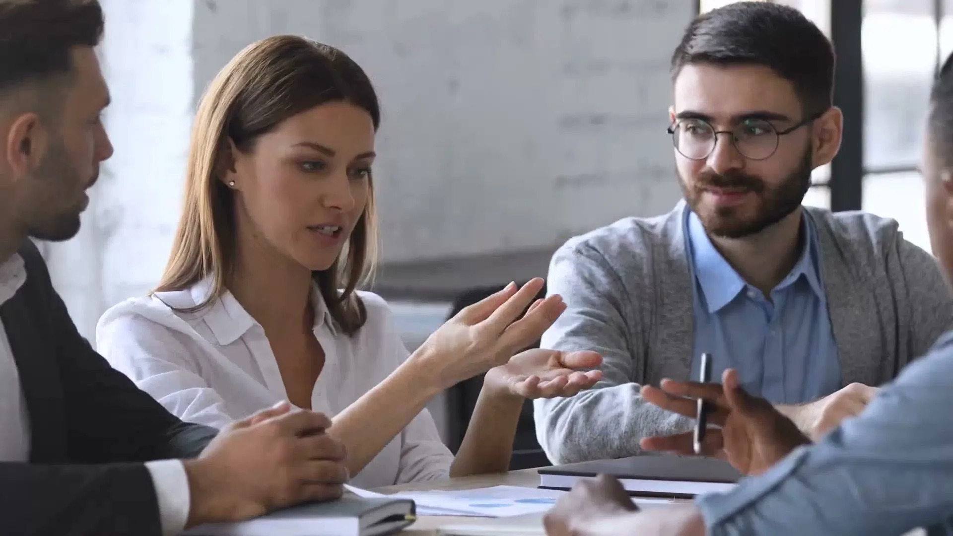 A diverse group of business professionals, including a woman in a white shirt, are engaged in a discussion around a table in an office setting.