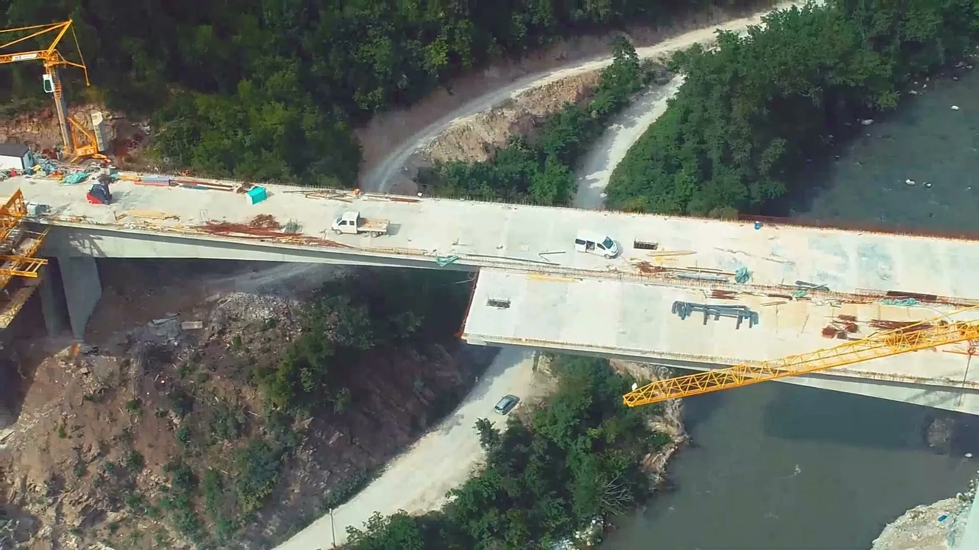 An aerial view shows a long concrete bridge under construction over a river, with cranes and vehicles on the unfinished deck, surrounded by green trees and dirt roads.
