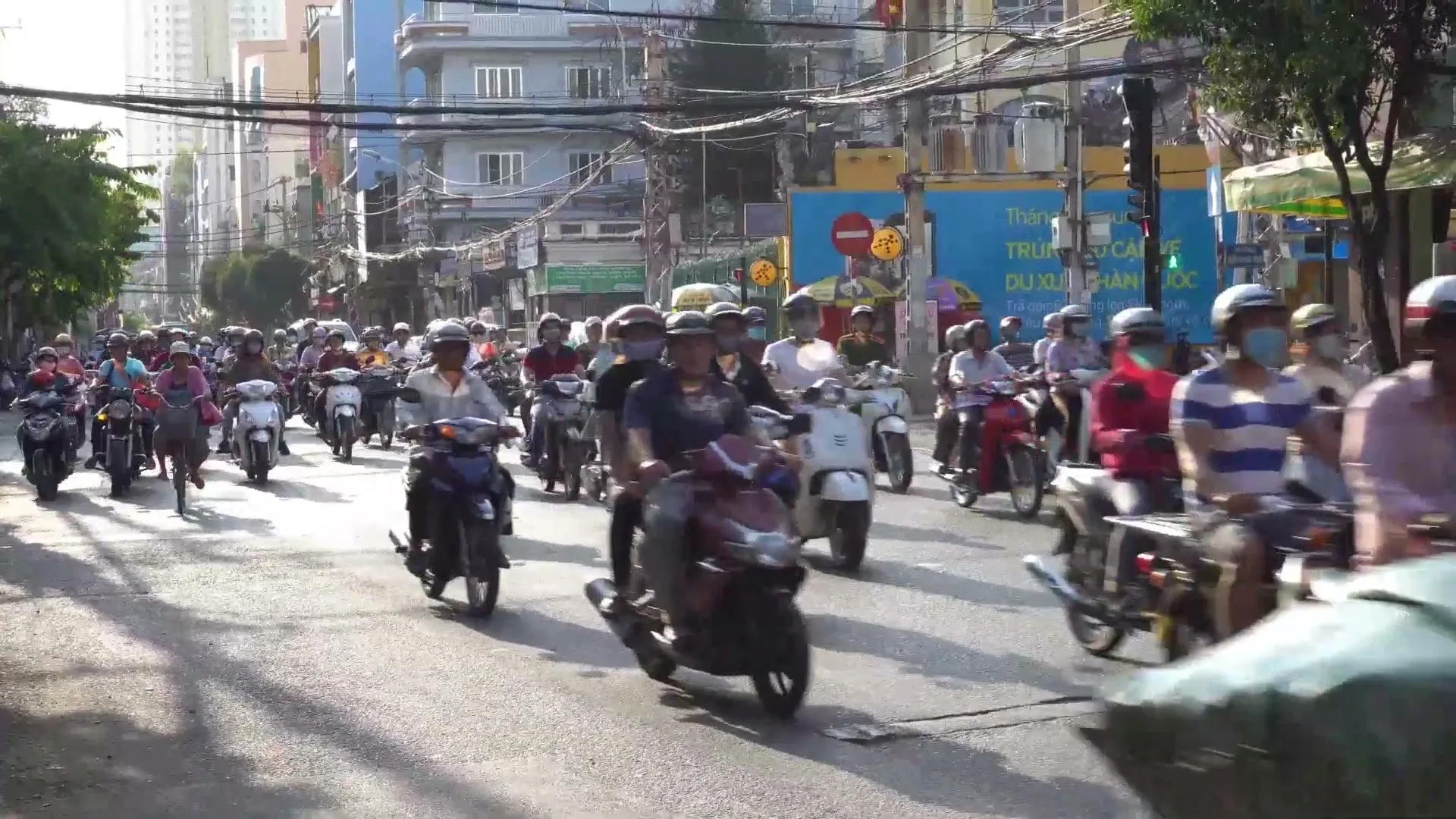 A wide shot captures a bustling street scene in a city, with numerous people on motorbikes filling the road, creating a sense of vibrant activity.