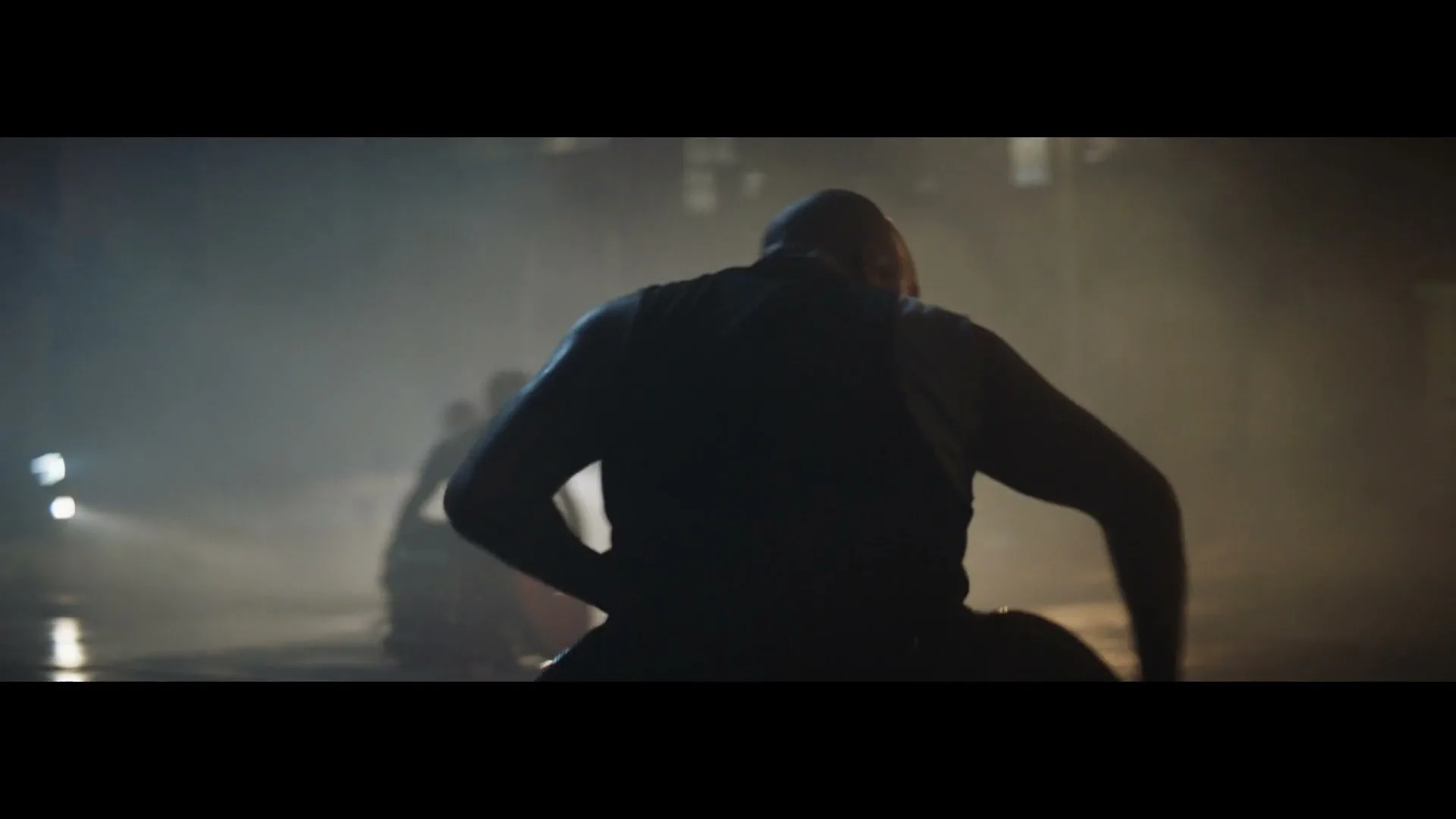 A male athlete in a tank top is seen from behind, silhouetted against a smoky, dimly lit background, preparing to run.