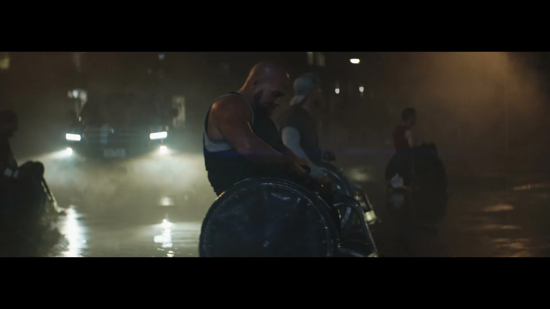 A male wheelchair athlete in a tank top looks down, exhausted, as other athletes and a car with headlights on are visible in the background on a wet street at night.