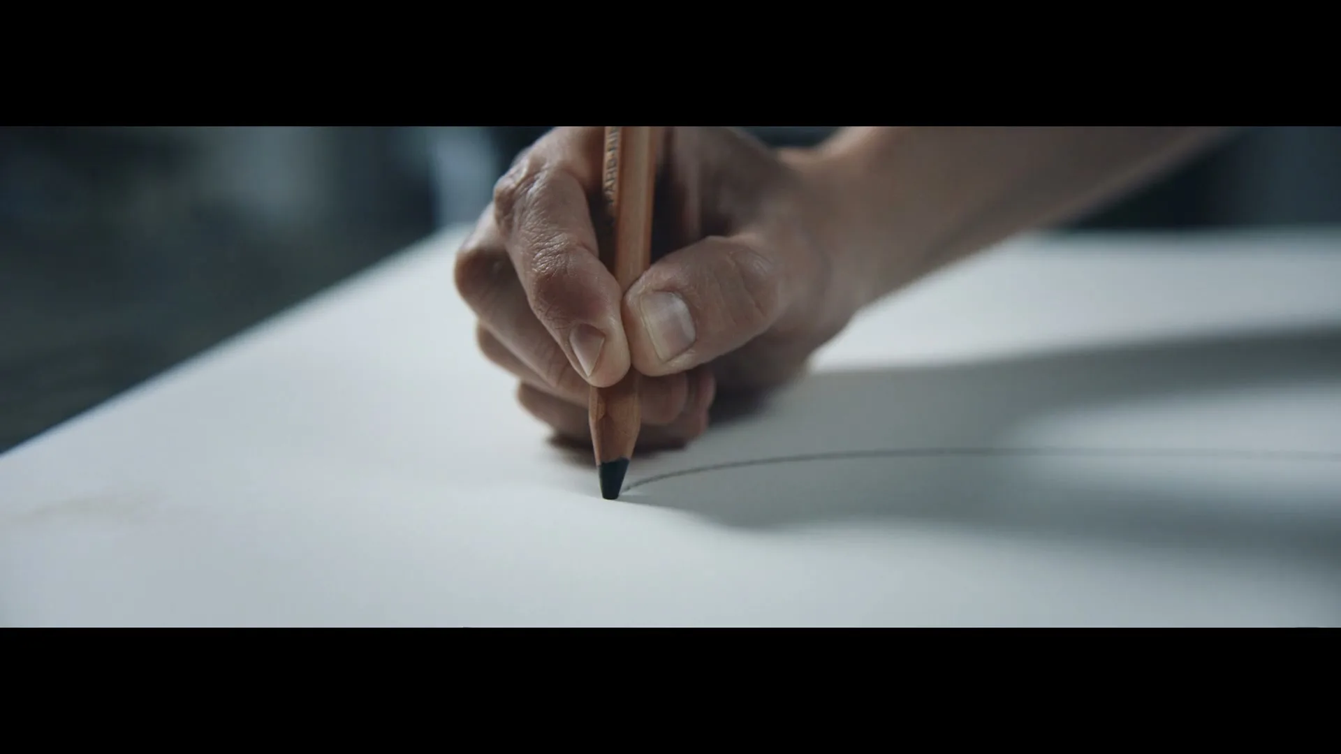 A person's hand holds a pencil, drawing a line on a white sheet of paper in an interior setting.