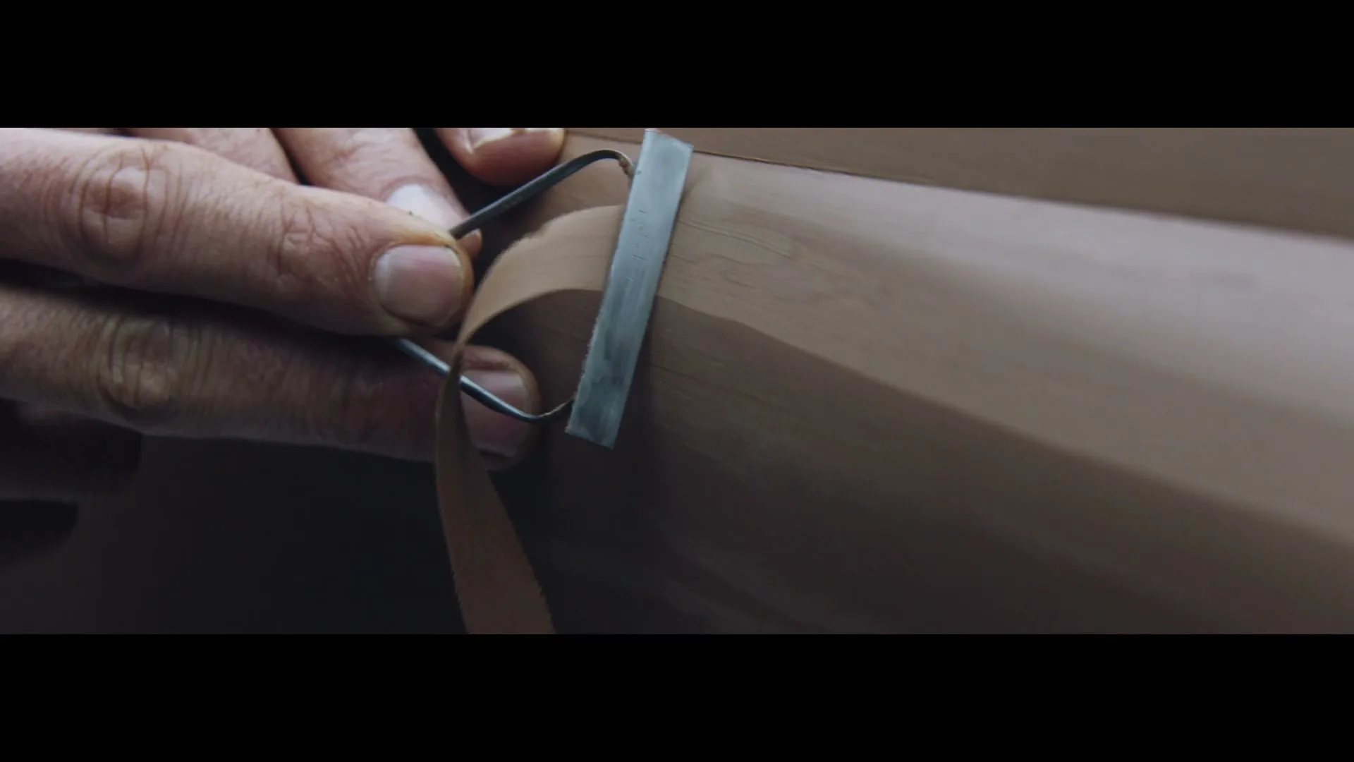 A person's hands meticulously sculpt a clay model with a wire tool, shaping a smooth, curved surface in a close-up shot.