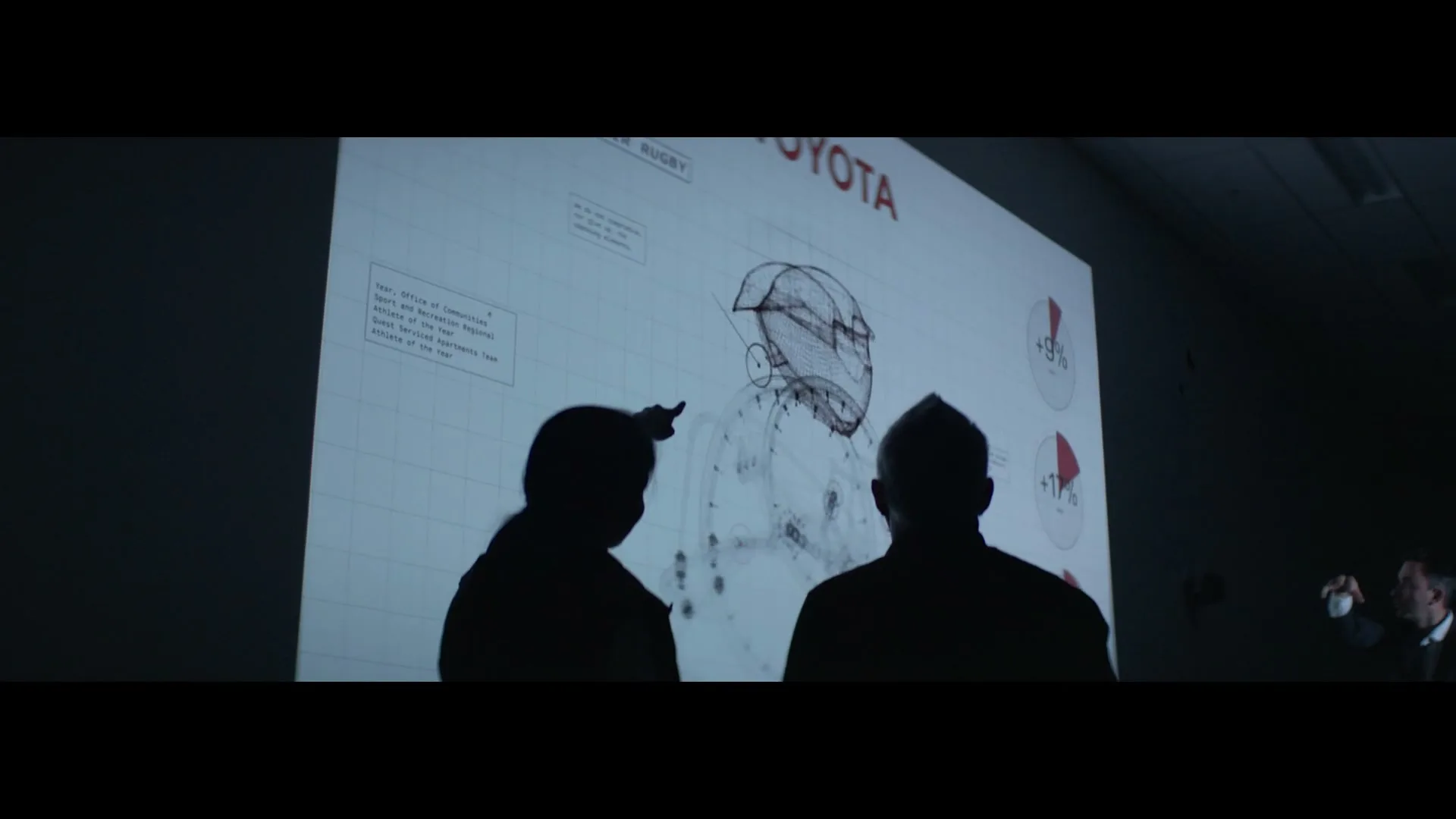 Two silhouetted people stand in a dark room, looking at a large projection of a technical drawing and text on a screen.