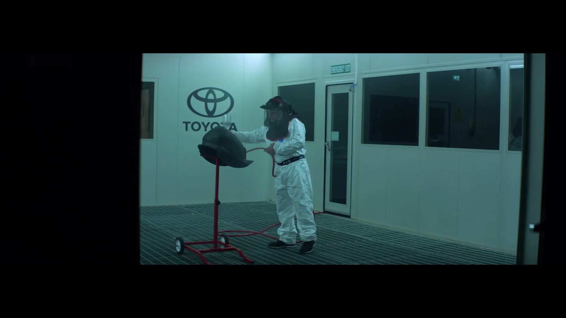 A person in a white hazmat suit and face shield sprays a car part on a stand in a brightly lit, sterile white room with a Toyota logo on the wall.