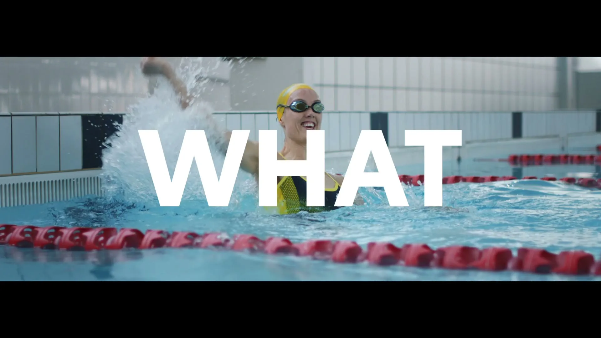 A female swimmer in a yellow cap and goggles splashes water in a pool, smiling with the word "WHAT" superimposed.