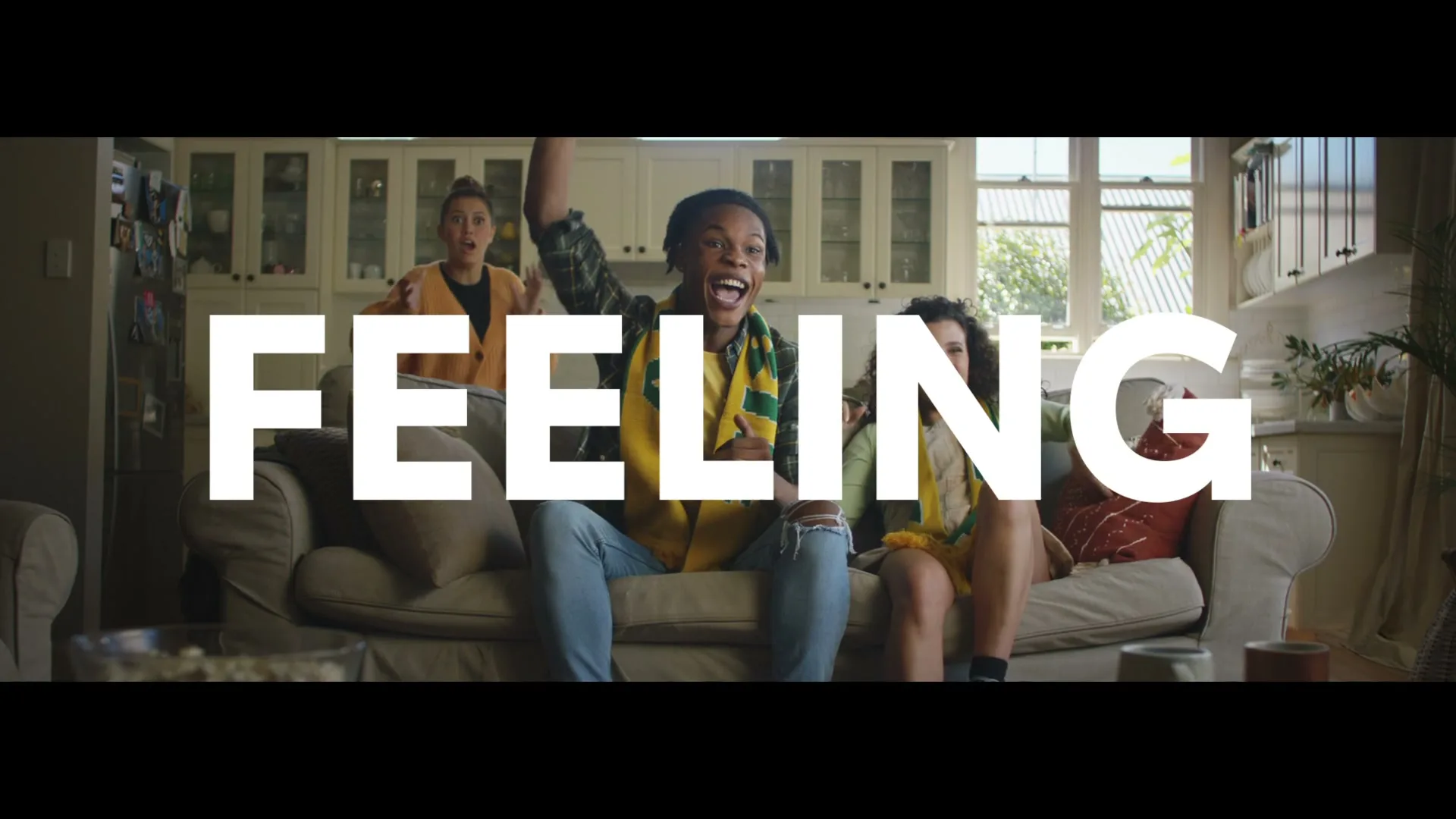 Three young adults, two females and one male, sit on a couch in a living room, cheering excitedly while watching something off-screen, with the word "FEELING" superimposed.