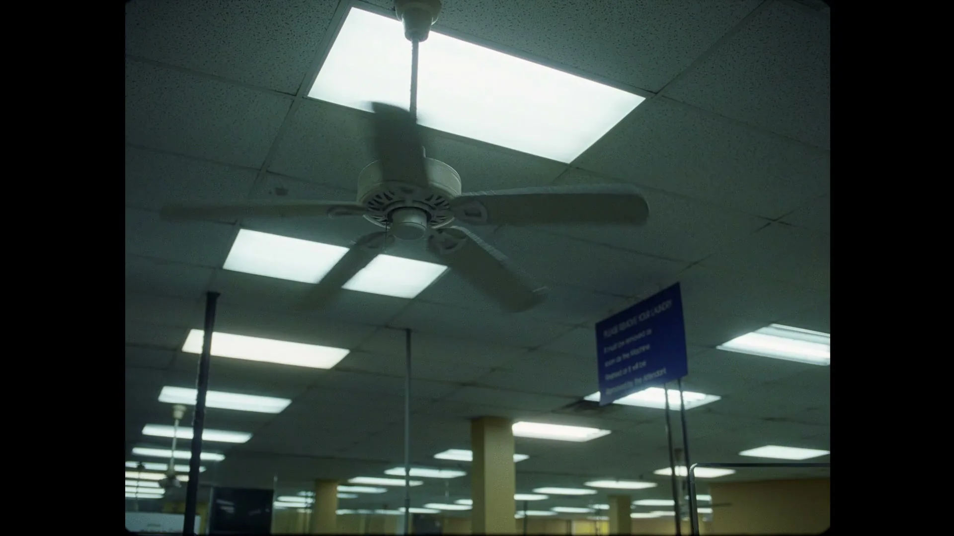 A low-angle shot of a white ceiling fan spinning slowly in a room with fluorescent lights and a blue sign.