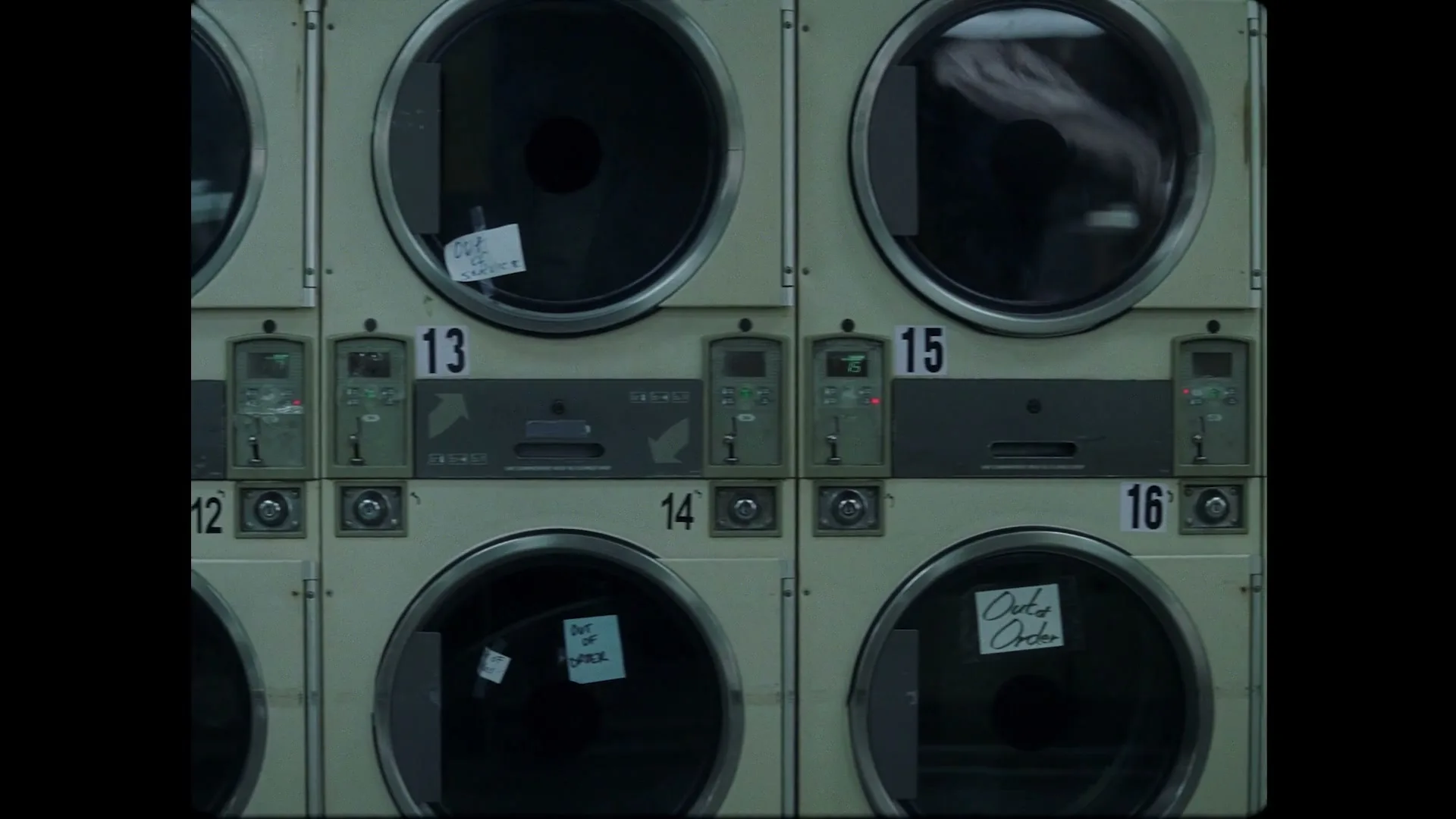 A grid of industrial washing machines and dryers, some with "Out of Order" signs, are seen in a laundromat.