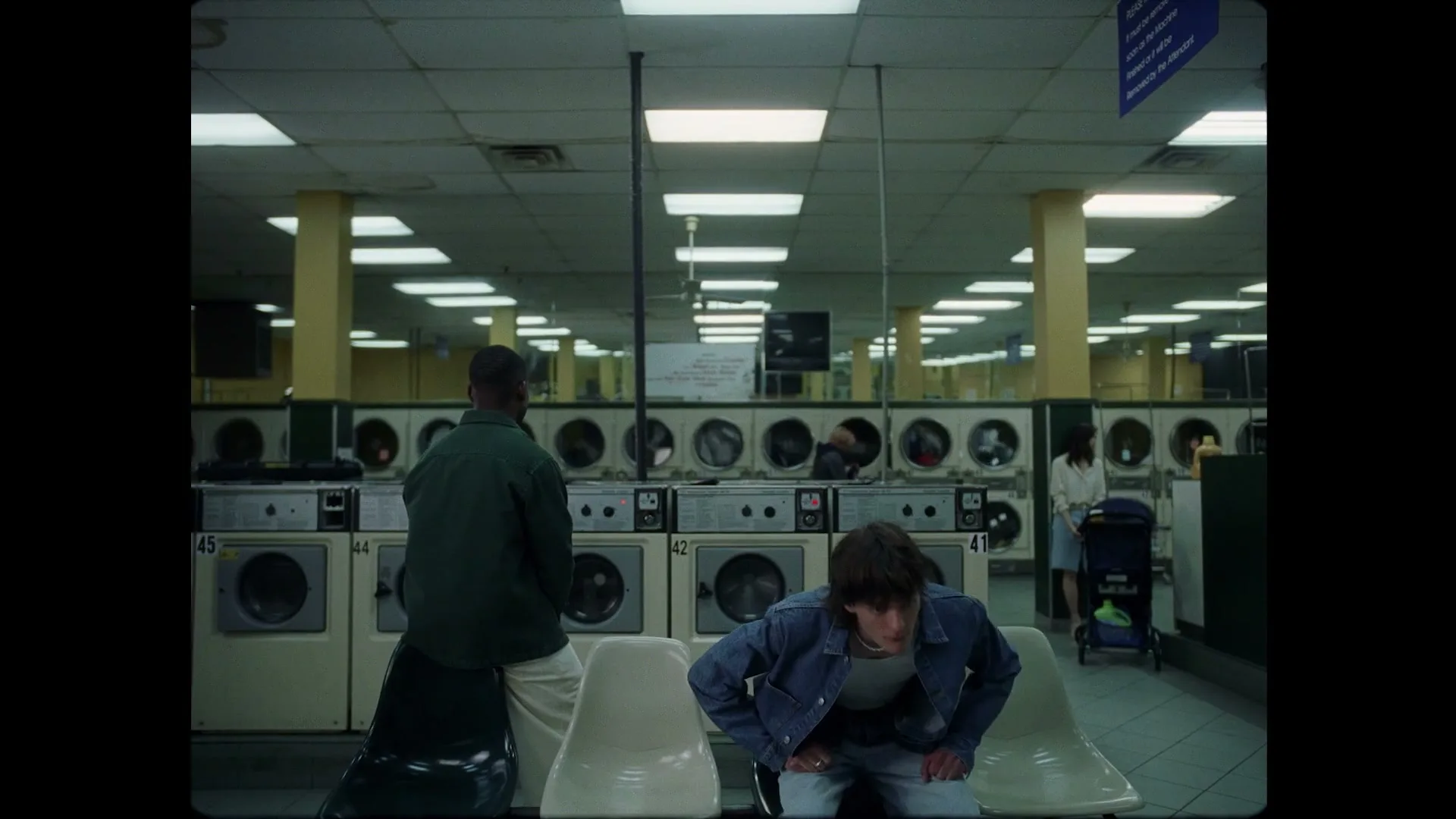 A young man with brown hair and a denim jacket walks into a laundromat, sits on a plastic chair, and looks around with a neutral expression.