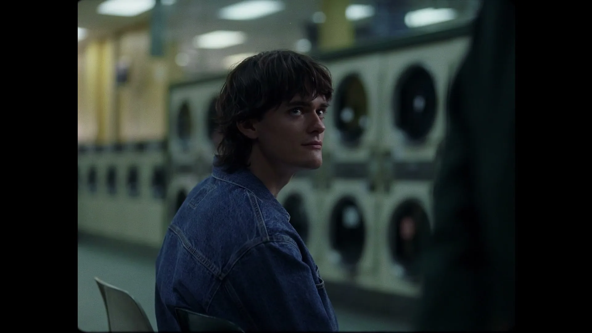 A young man with dark hair and a denim jacket sits in a laundromat, looking to his right as he talks to someone off-screen.