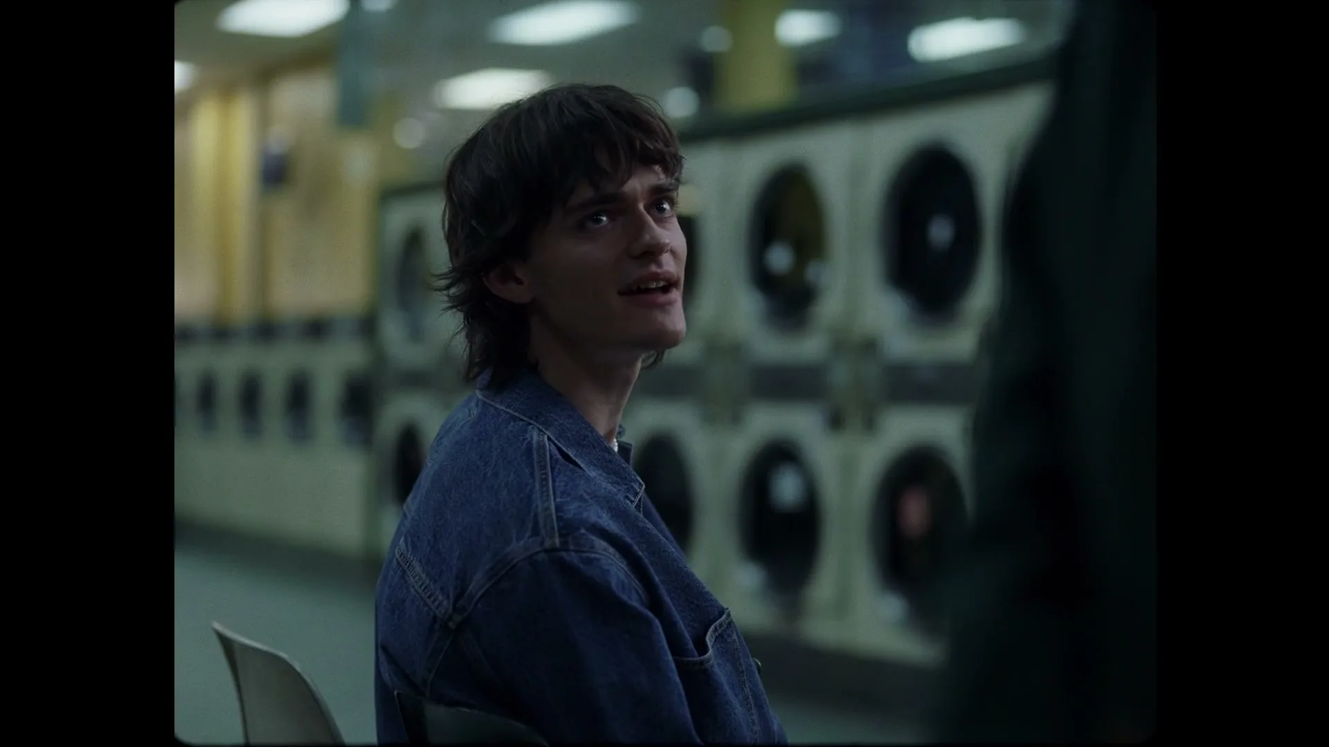 A young man with dark hair and a denim jacket sits in a laundromat, looking up and to the right with a slight smile.