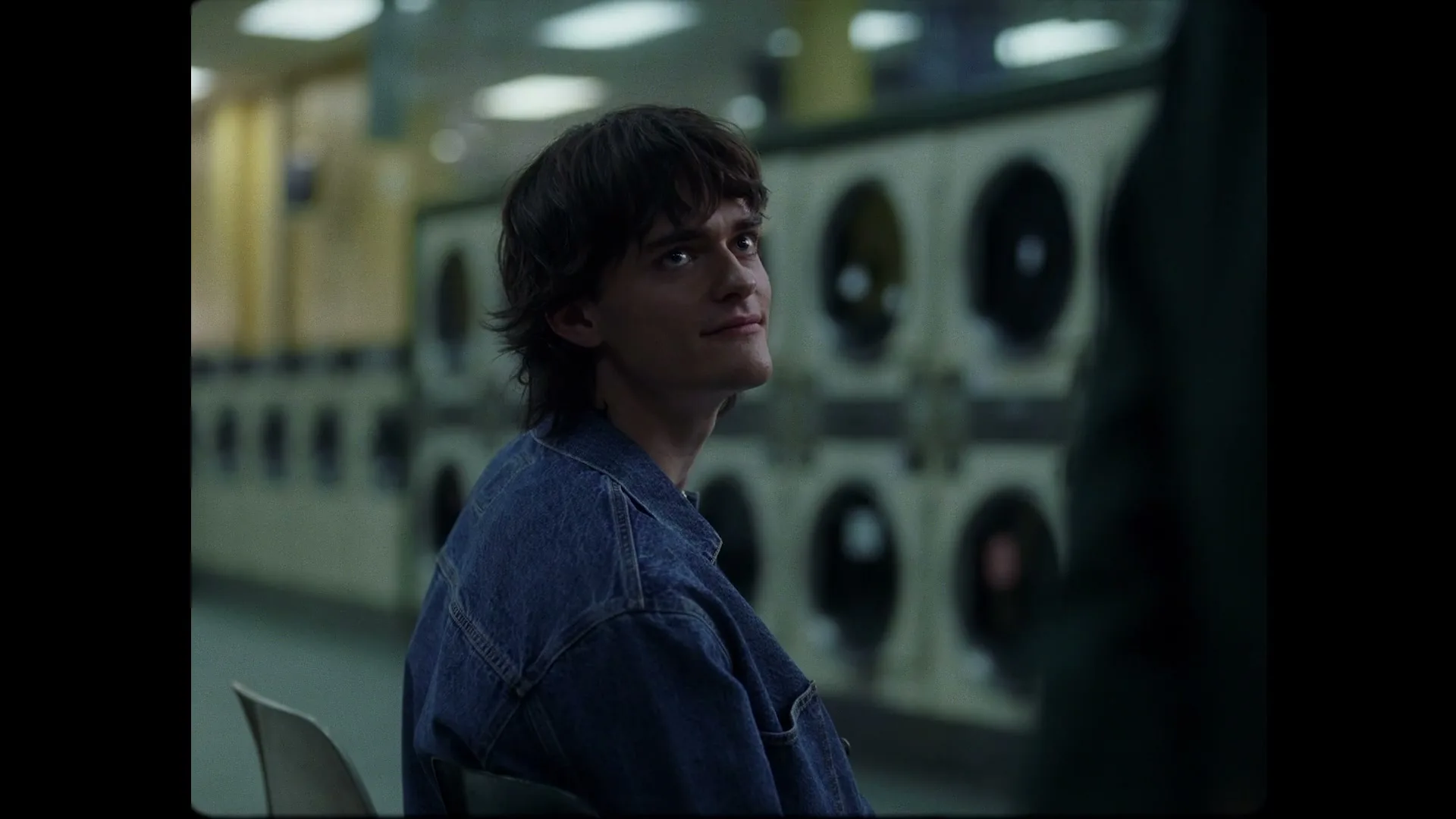 A young man with dark hair and a denim jacket sits in a laundromat, looking up and smiling slightly.