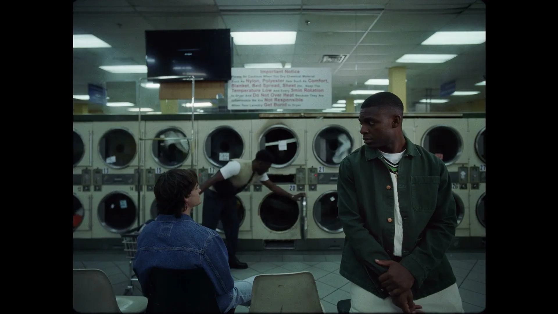 Two men sit in a laundromat while another man loads a washing machine in the background.