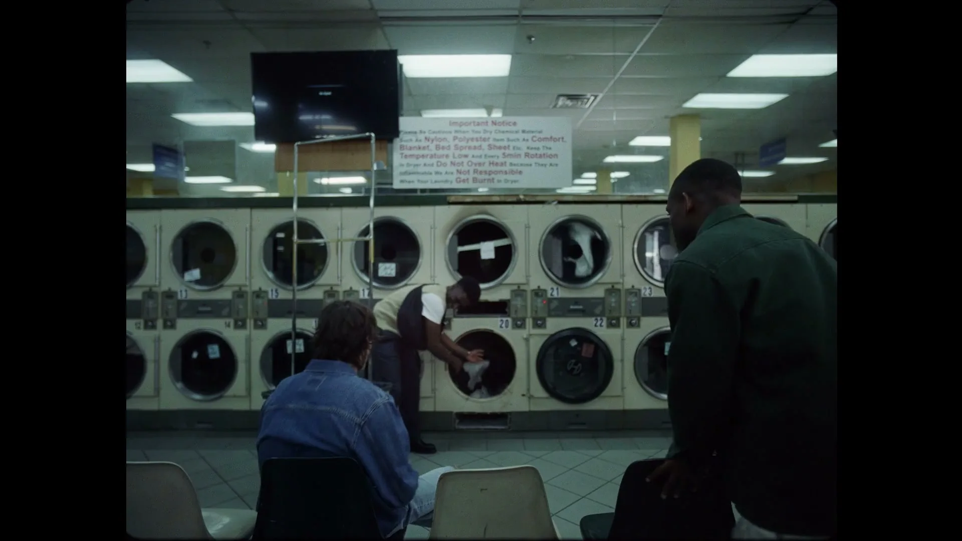 Three young men in a laundromat are surprised by something in a washing machine, then they all gather around it to investigate.