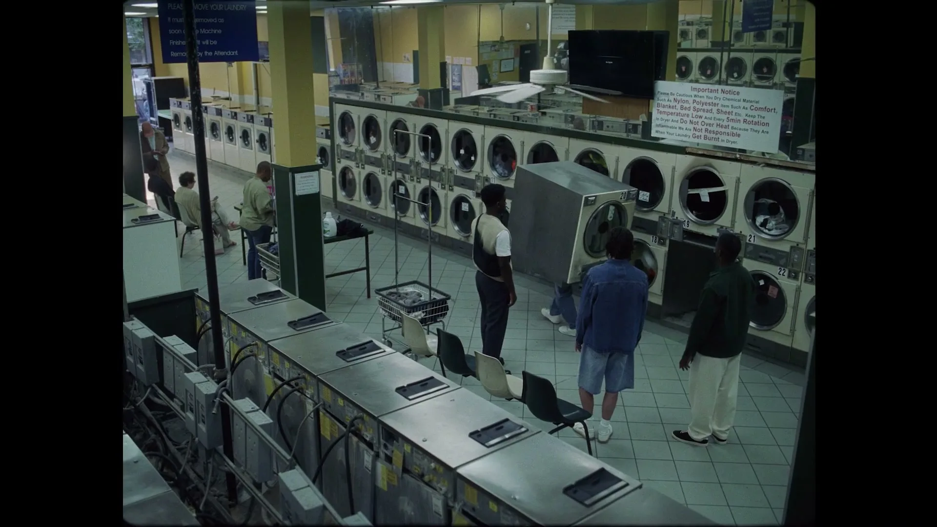 A group of young men are in a laundromat, with one man carrying a large washing machine, as others watch him in a wide shot.