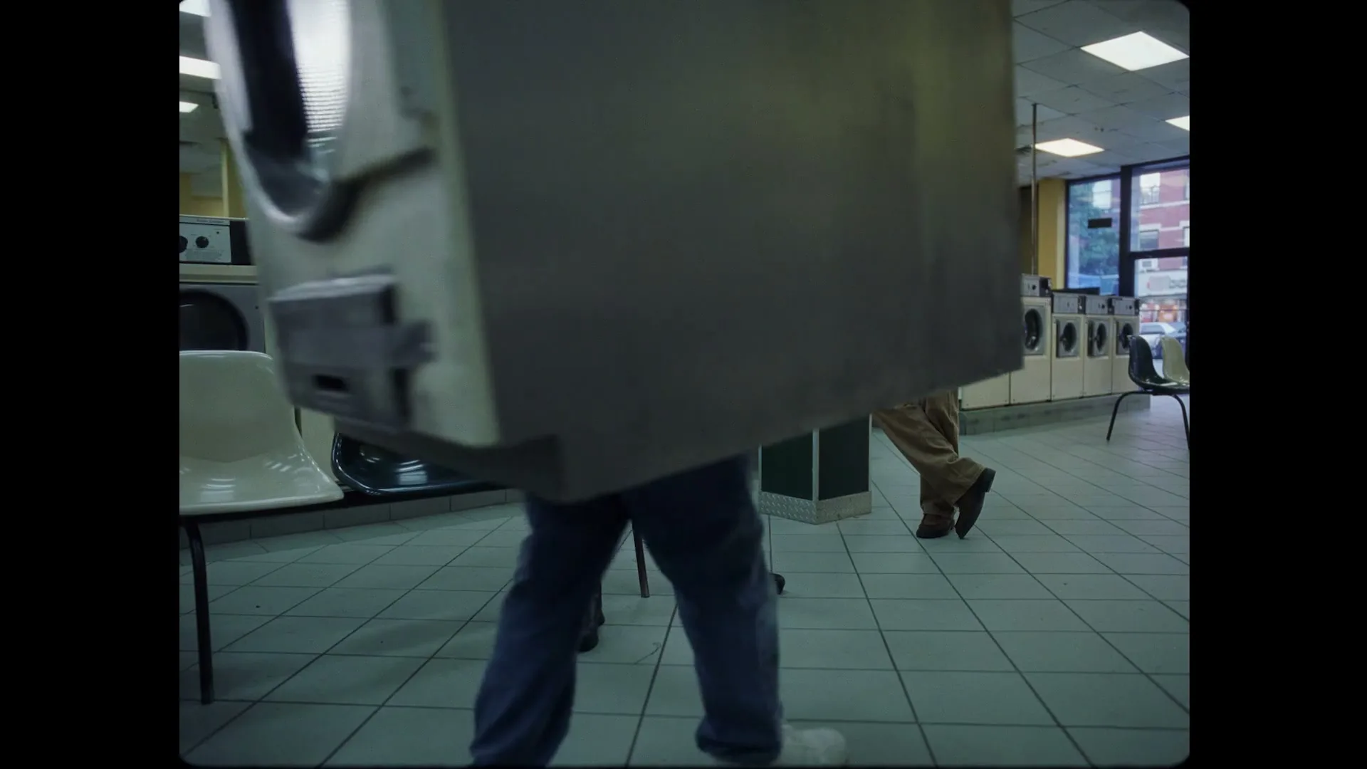 A man in black shorts and jacket sits on a chair in a laundromat, while another man in a tan suit leans against a pillar, and a third person walks by carrying a washing machine.