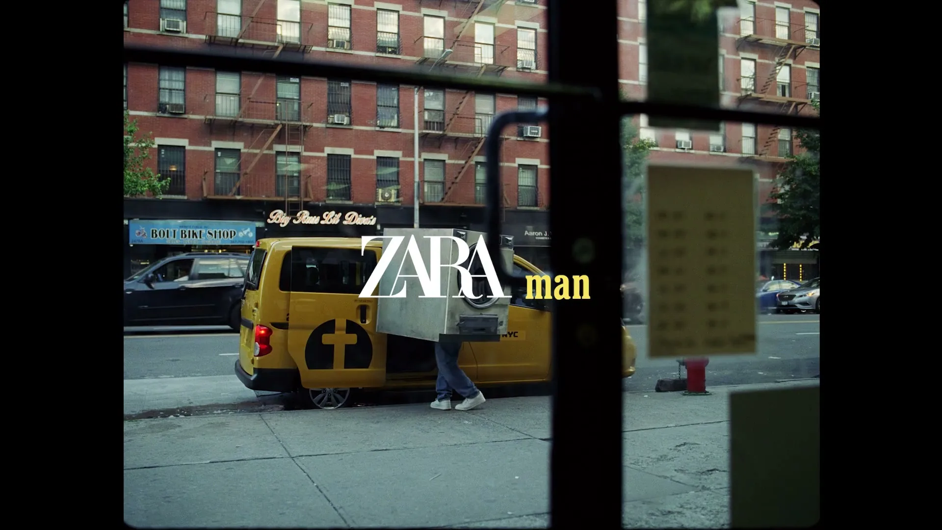 A man with a washing machine on his back walks down a city street, as the camera tracks him from inside a building.