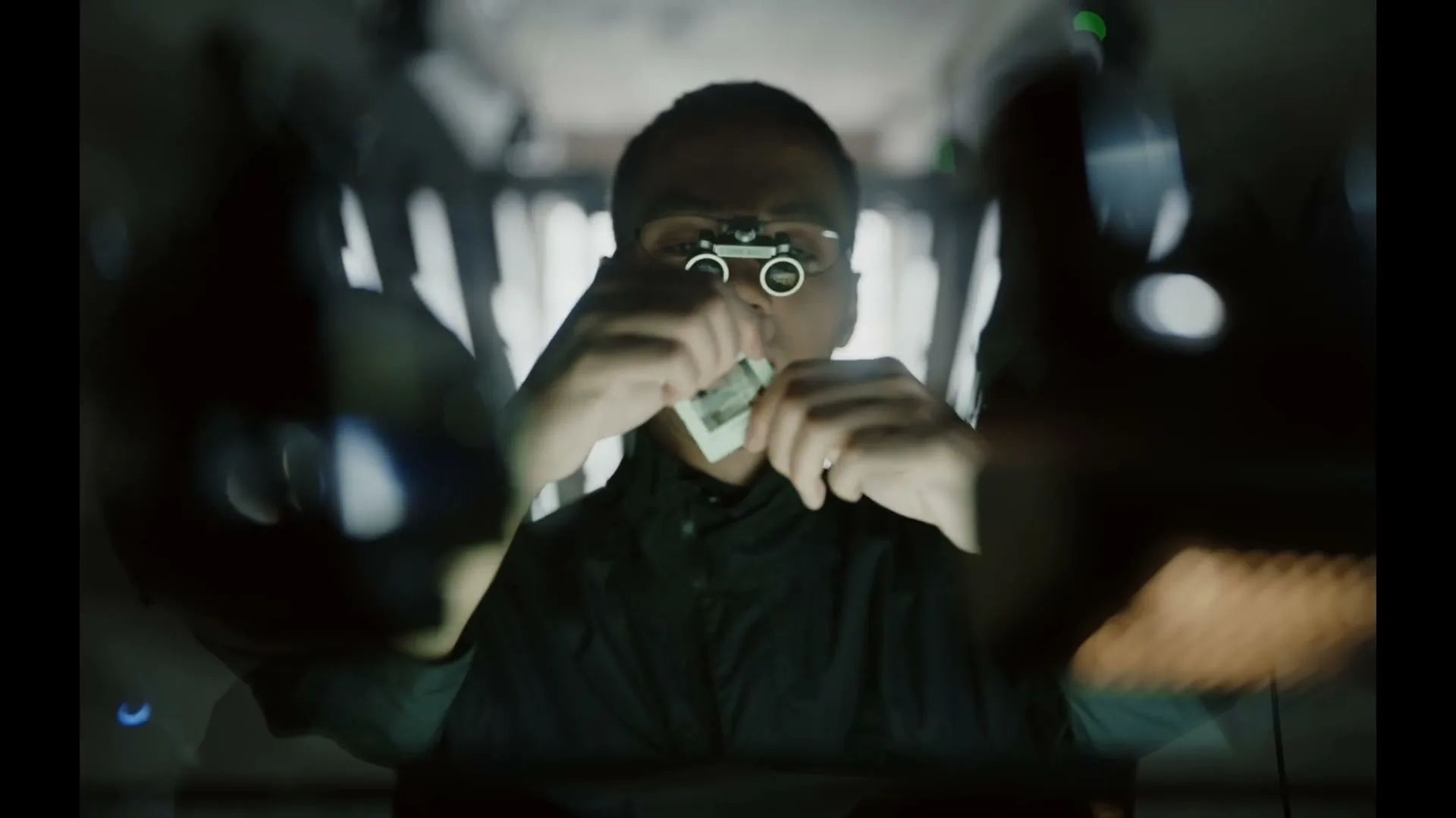 A man wearing magnifying glasses examines a photographic slide, holding it up to the light in a dark, industrial-looking interior.