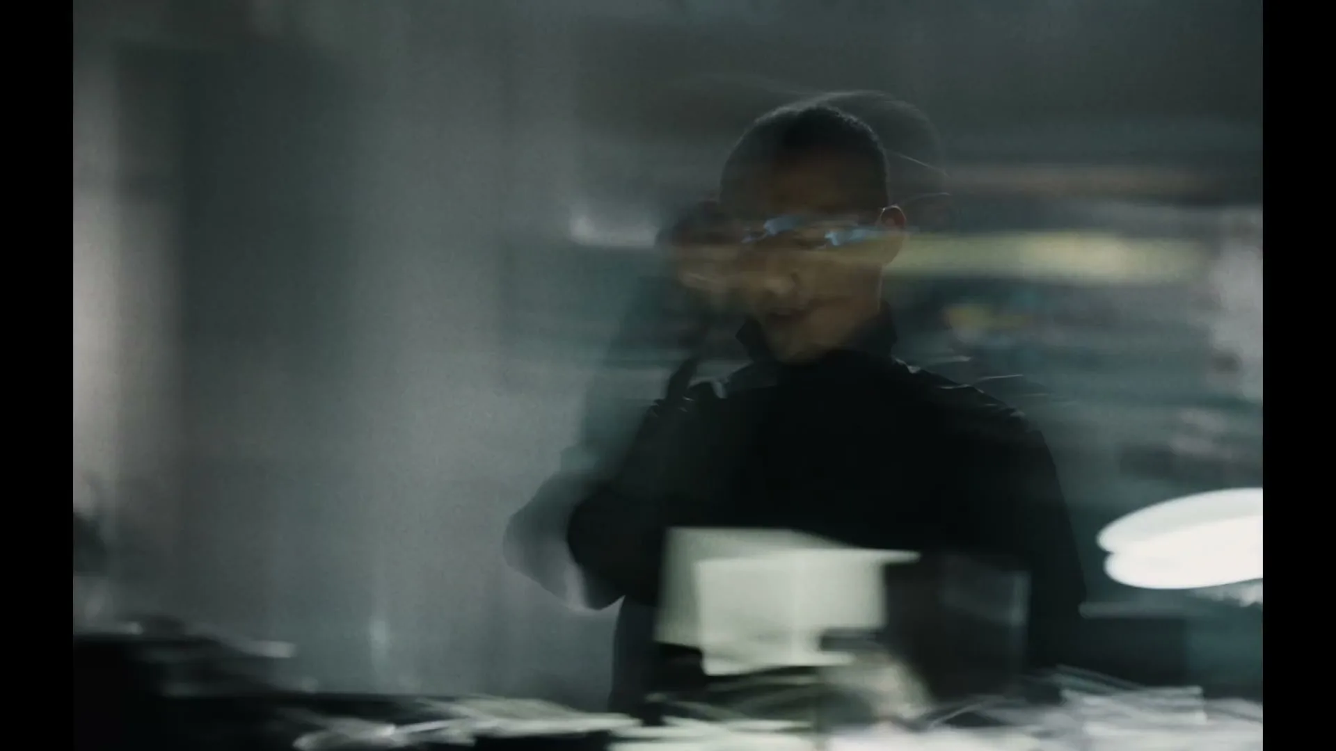 A man in glasses and a dark shirt is blurred, moving quickly in a dimly lit, industrial-looking interior space.