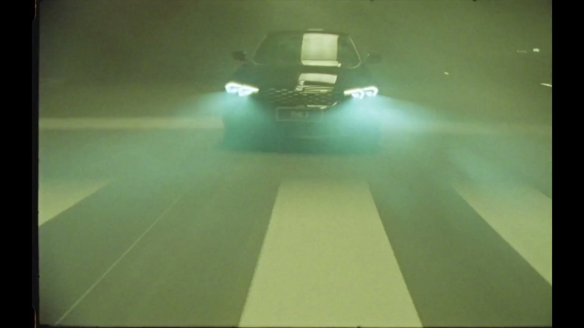 A dark BMW car with bright headlights drives towards the camera through a smoky, green-tinted tunnel, revealing road markings.