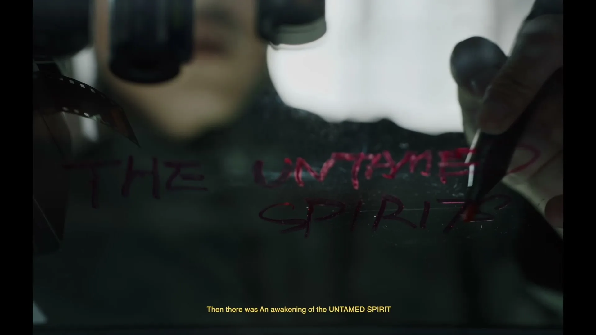 A person's hand writes "THE UNTAMED SPIRIT" in red on a glass surface, with film reels visible in the background.