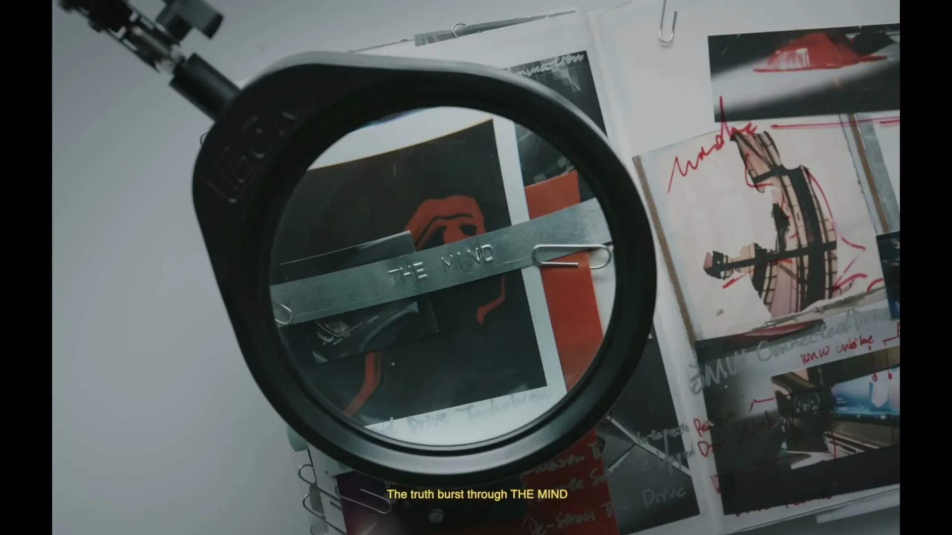 A magnifying glass reveals a metal tag with the words "THE MIND" over a notebook with sketches and notes, on a white surface.
