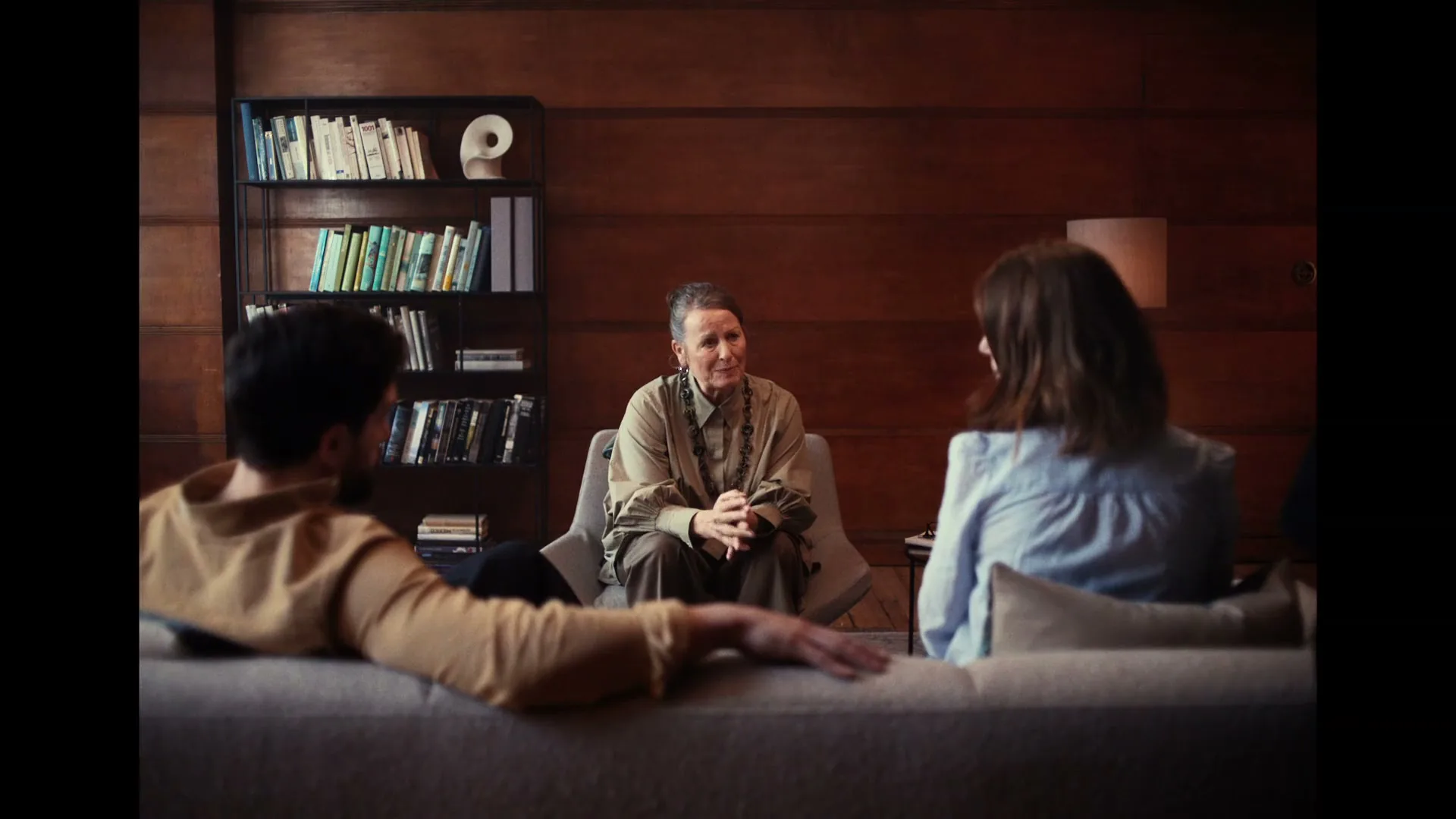 A therapist sits between a couple on a couch, talking to them in a wood-paneled office.