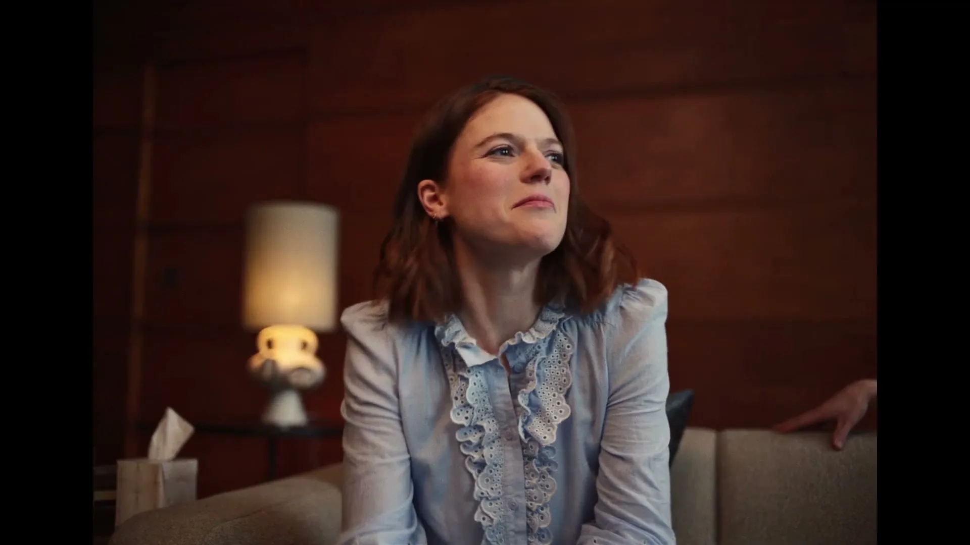 A young woman with brown hair, wearing a light blue ruffled shirt, sits on a couch in a wood-paneled room, looking around with a slightly annoyed expression.