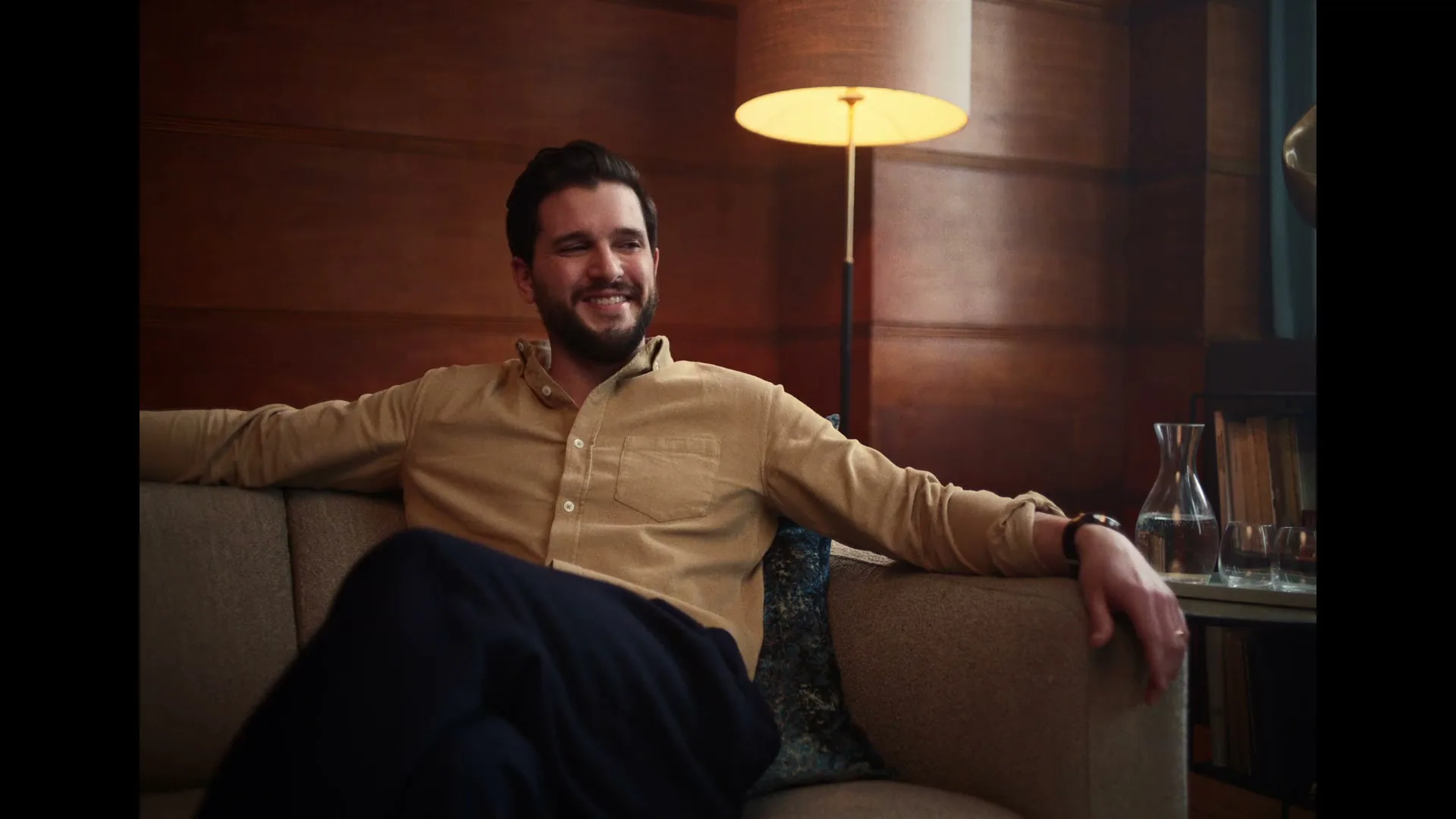 A man with a beard, wearing a tan shirt and dark pants, sits comfortably on a sofa in a warmly lit room, smiling slightly.