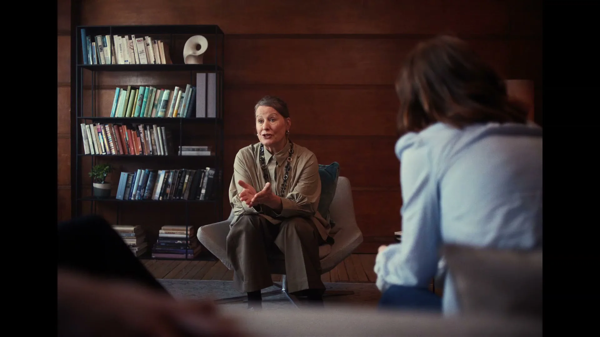 A female therapist in her 50s or 60s, wearing a beige shirt and dark pants, sits in a modern chair, gesturing with her hands while talking to a patient in a wood-paneled office.