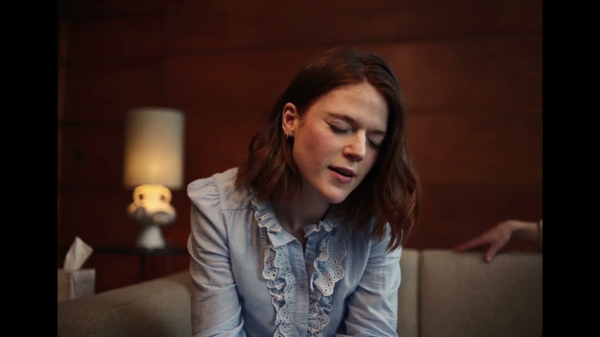 A woman with short brown hair and a light blue ruffled shirt sits on a couch, looking down and speaking softly in a dimly lit room.