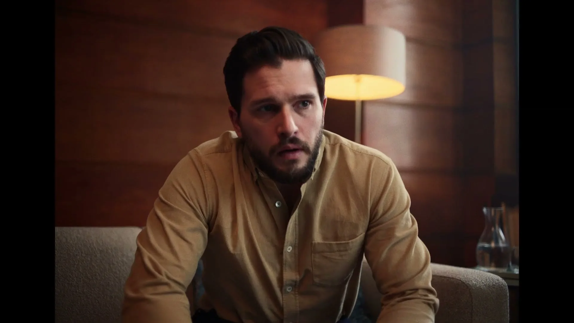 A man with a beard, wearing a tan shirt, sits on a couch in a dimly lit room, looking surprised and slightly confused.