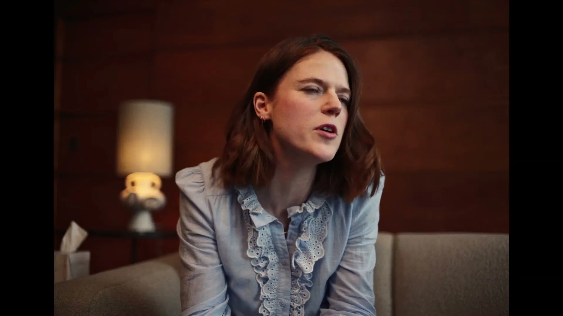 A young woman with brown hair, wearing a blue ruffled shirt, sits on a couch and talks with her eyes closed in a dimly lit room.