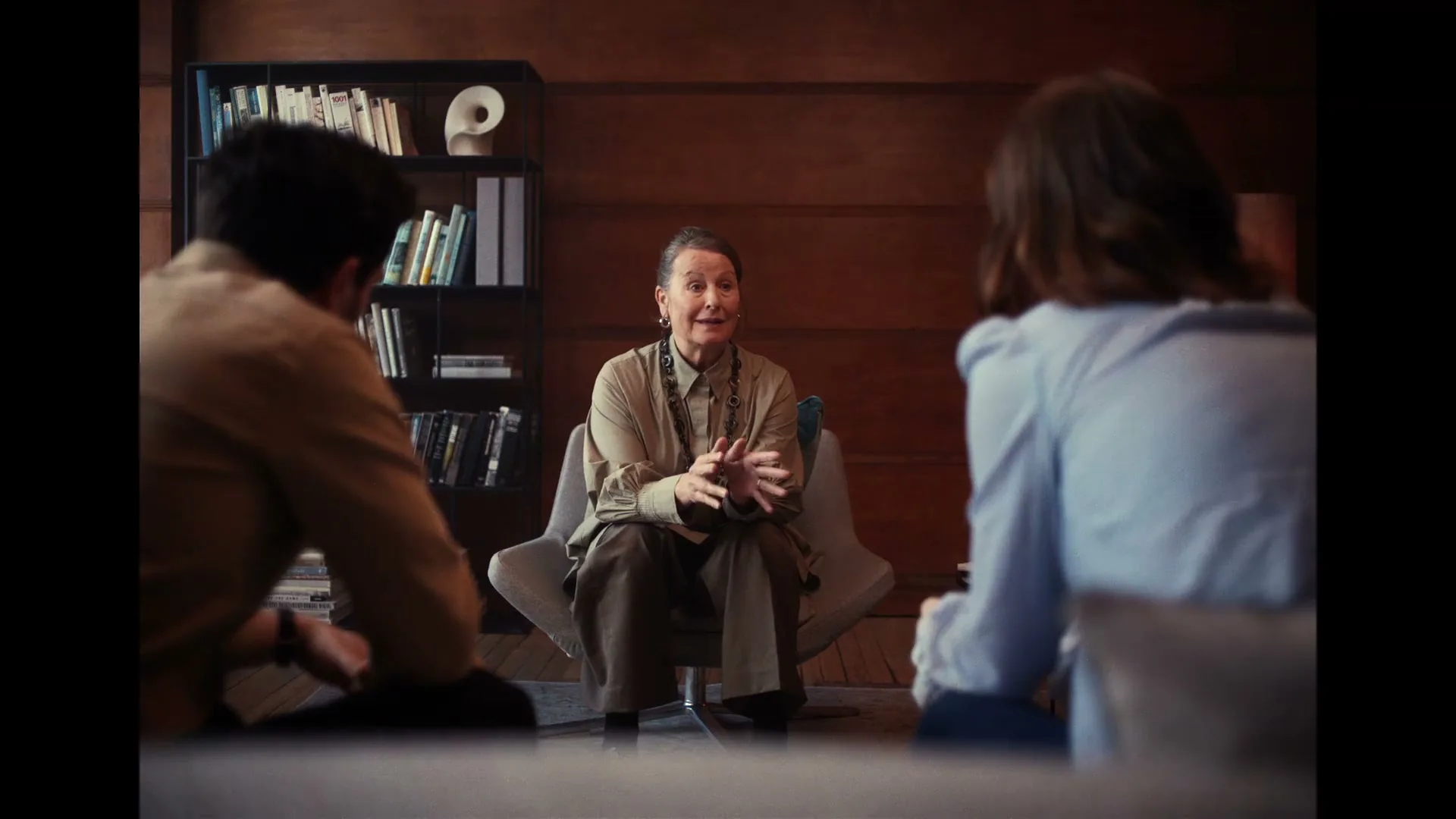An older female therapist in a beige outfit sits in a chair, gesturing with her hands, talking to a male and female couple in a modern office.