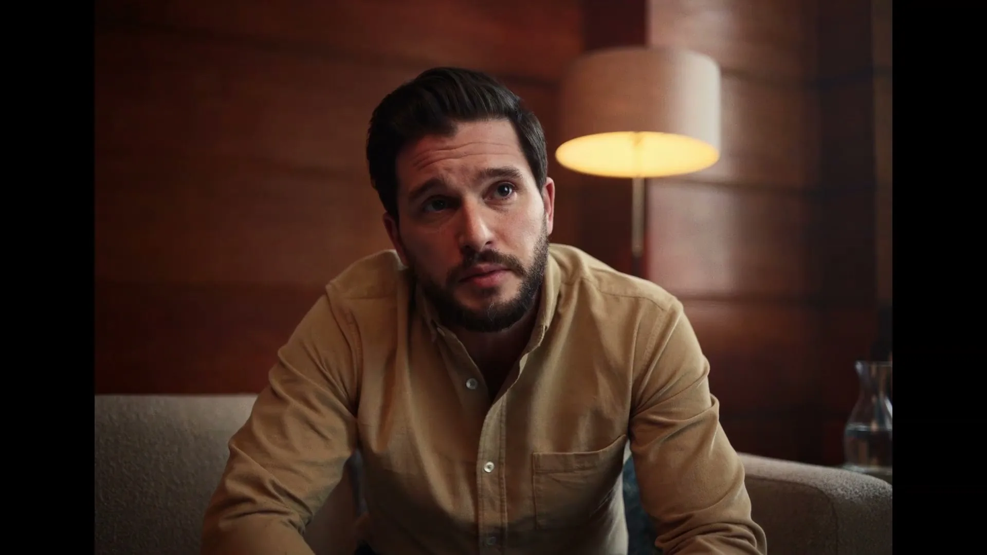 A man with a beard, wearing a tan shirt, sits on a couch in a dimly lit room, looking thoughtful and speaking.